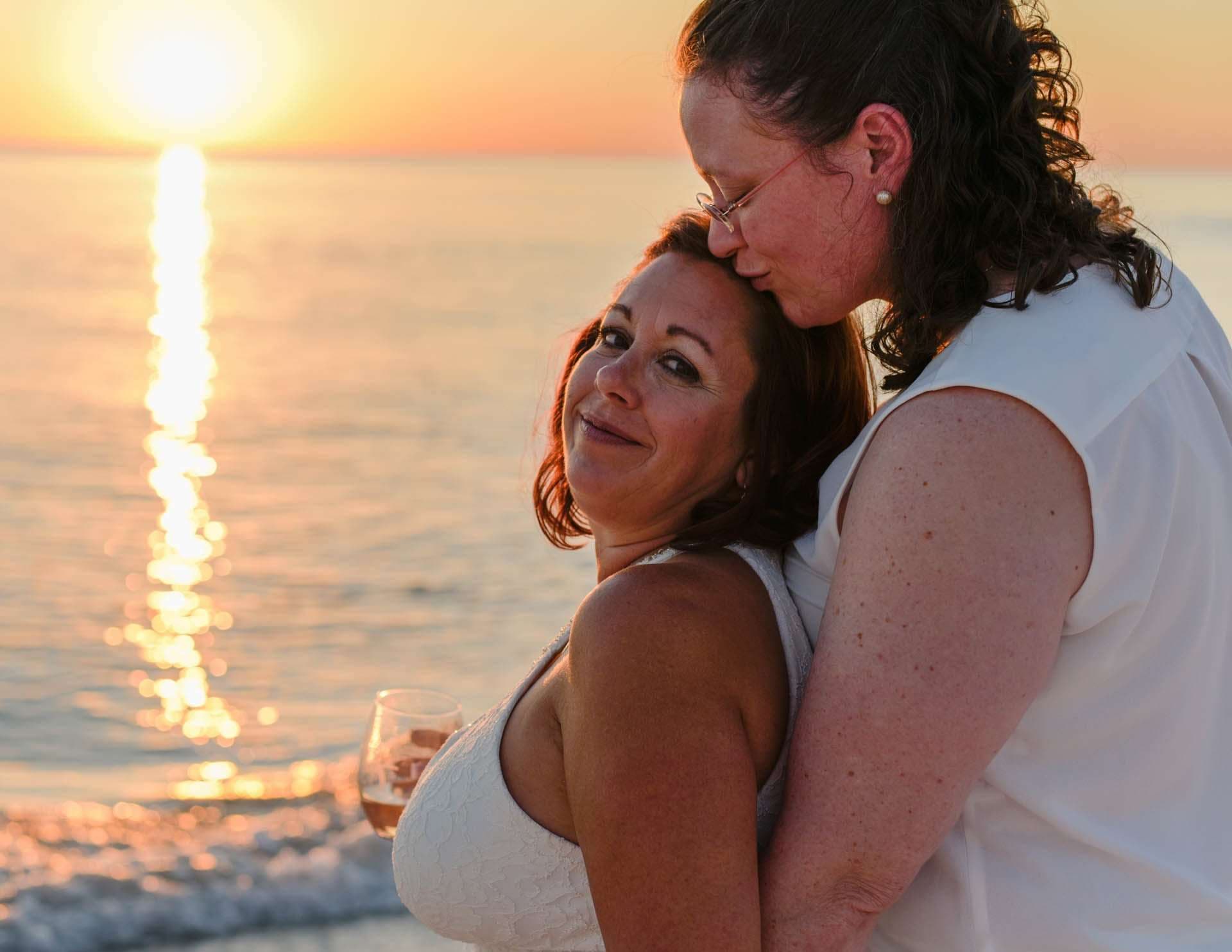 Two women in white embracing at sunset on the beach, one kissing the other's forehead as she holds a wine glass