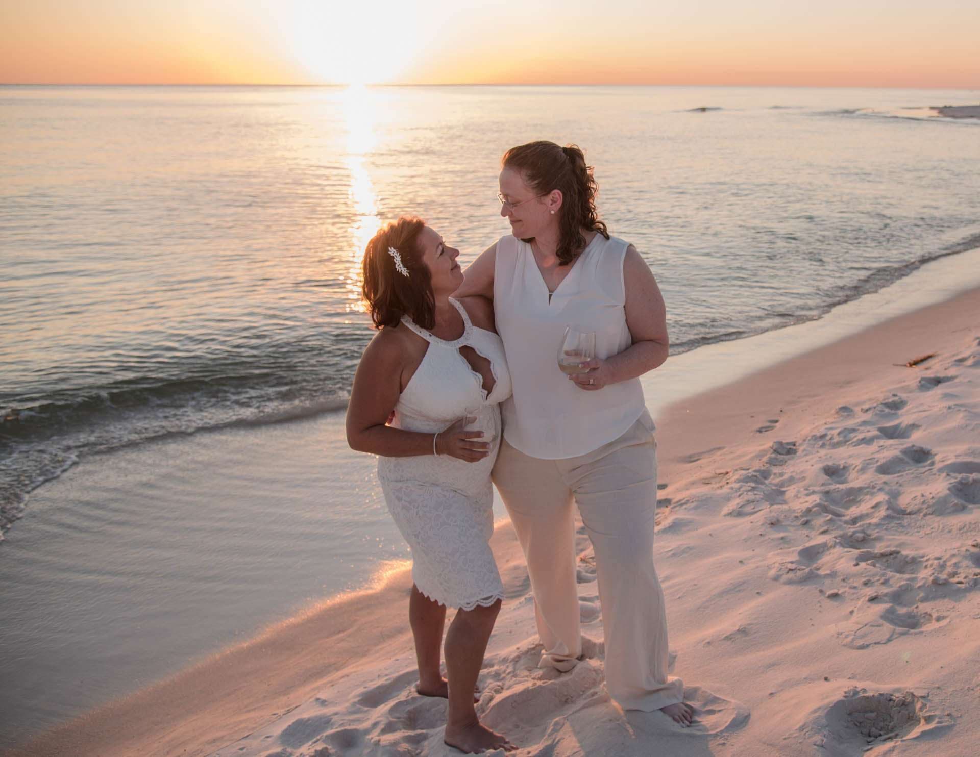 Two women in white embracing and smiling at each other on the beach at sunset, one holding a wine glass
