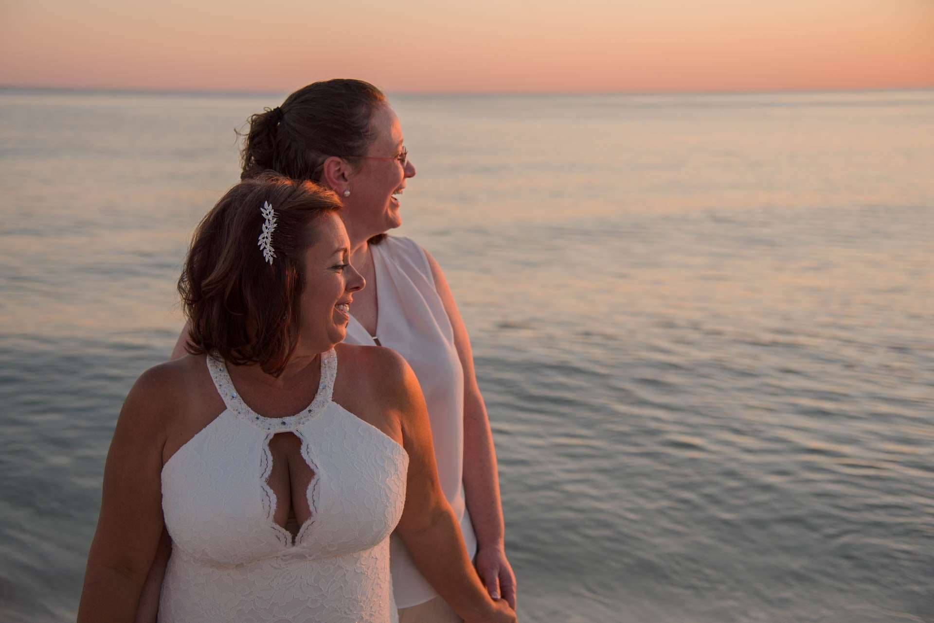 Two women in white laughing together by the water at sunset, one in a halter lace gown with a jeweled hair clip