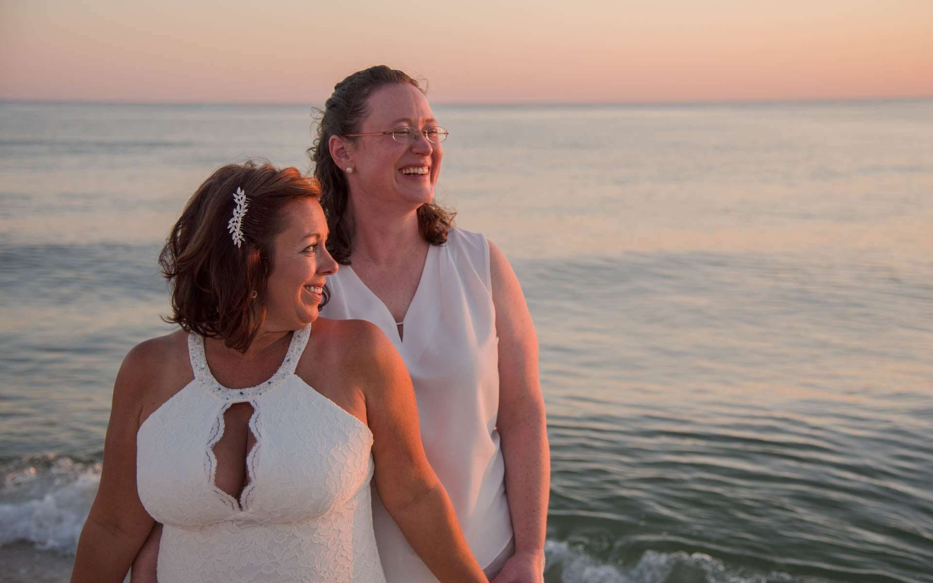 Two women in white laughing and looking out over the water at sunset on the beach