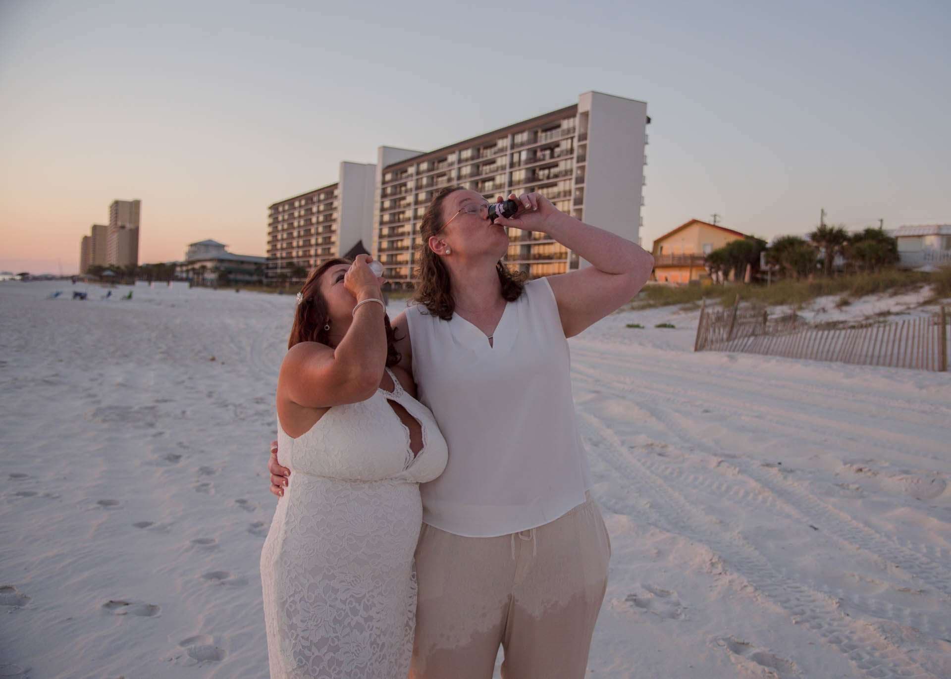Two women in white outfits embracing on the beach at sunset, each tipping back a small drink, condos behind them