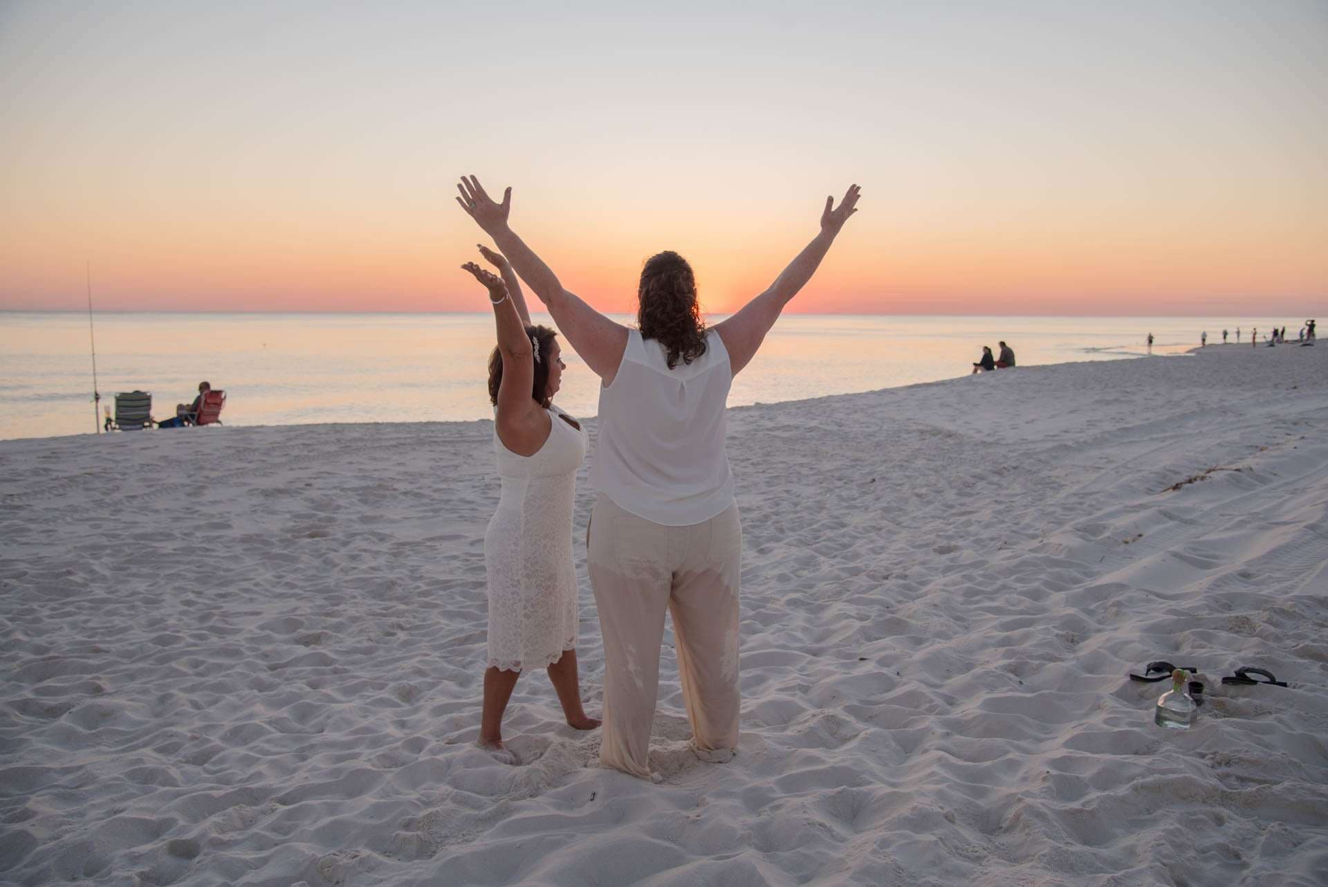 Two women in white raising their arms toward the sunset on a sandy beach at dusk