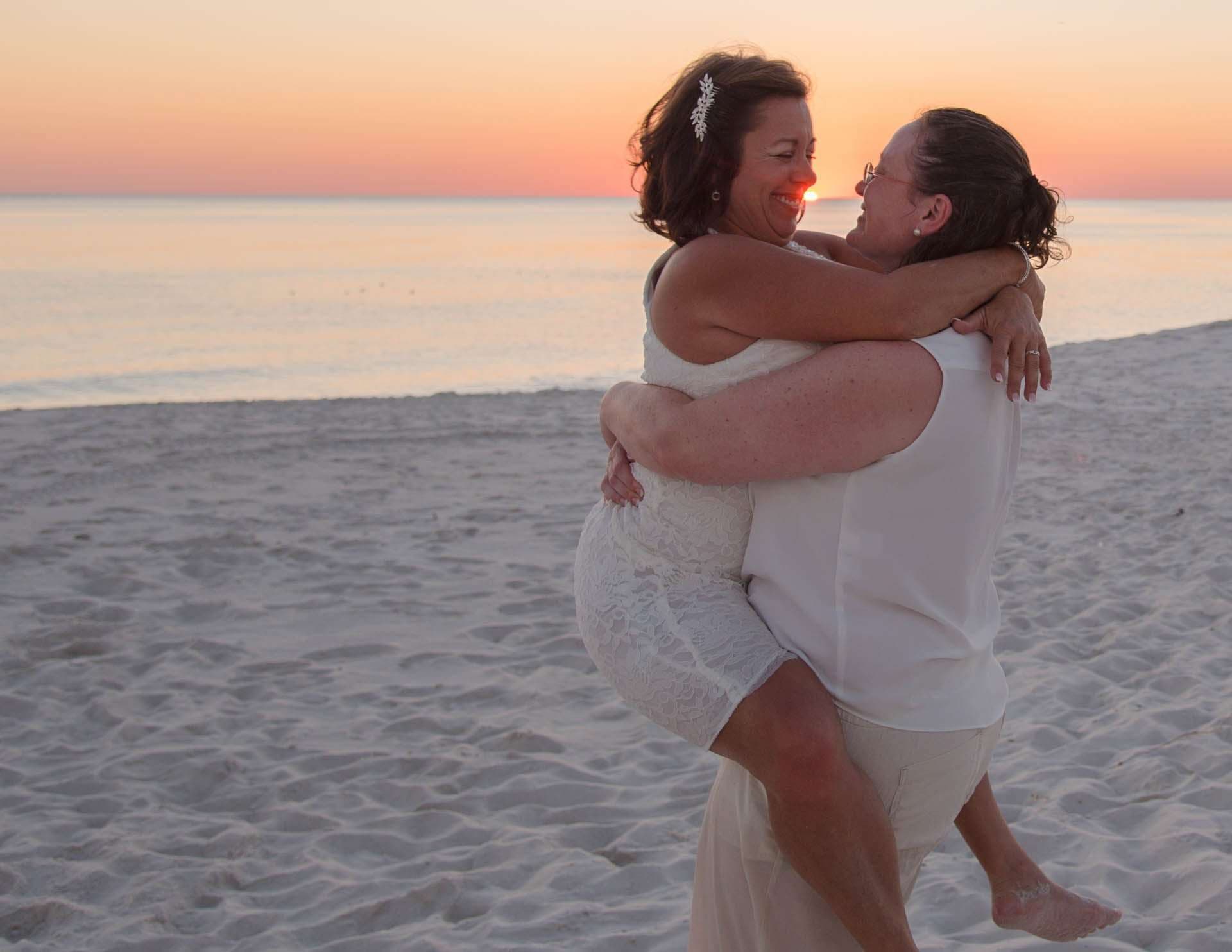 Two women in white embracing on the beach at sunset, one lifted off the ground, both smiling