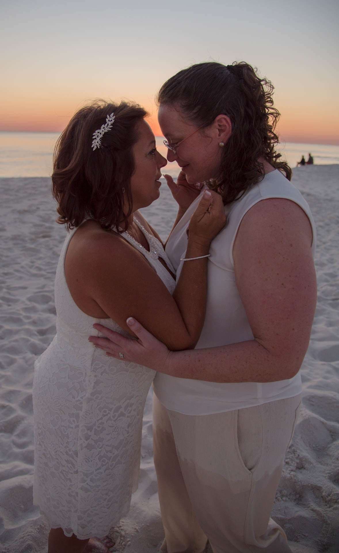 Two women in white embracing forehead to forehead on the beach at sunset, one wearing a sparkly hair clip