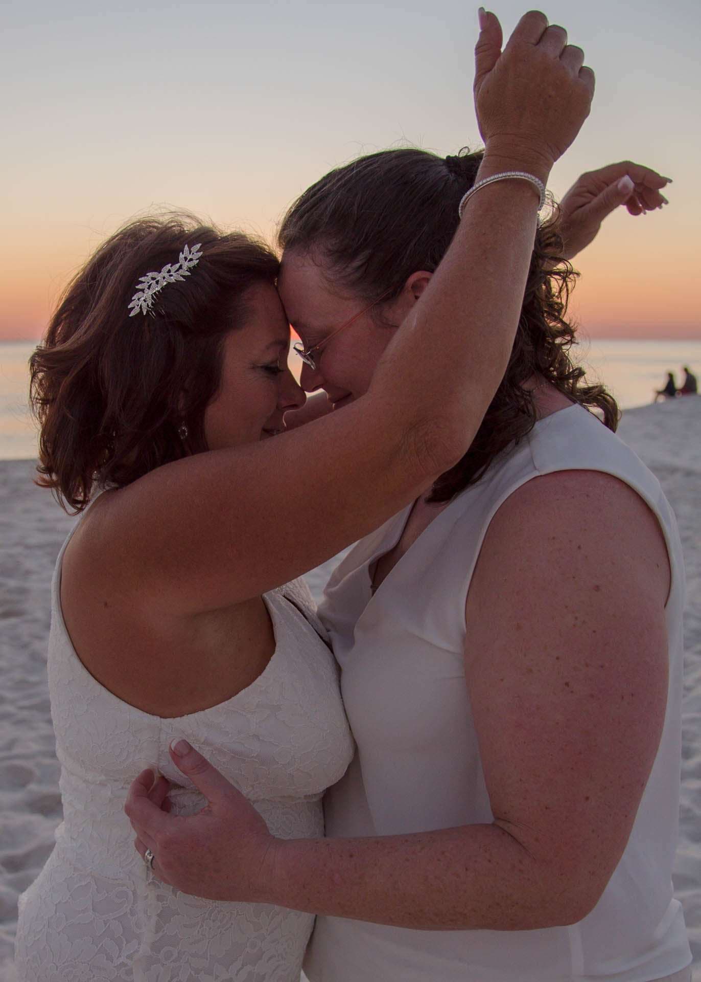 Two women in white touching foreheads and embracing on the beach at sunset, one wearing a sparkling hair clip