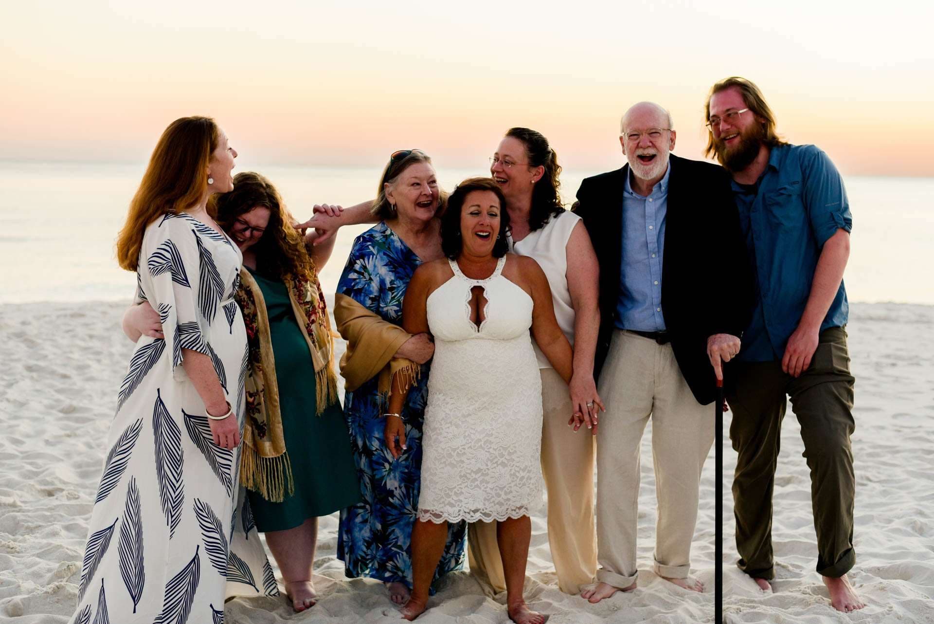 Seven laughing family and friends embrace in a row on the beach at sunset, the woman in white lace at center