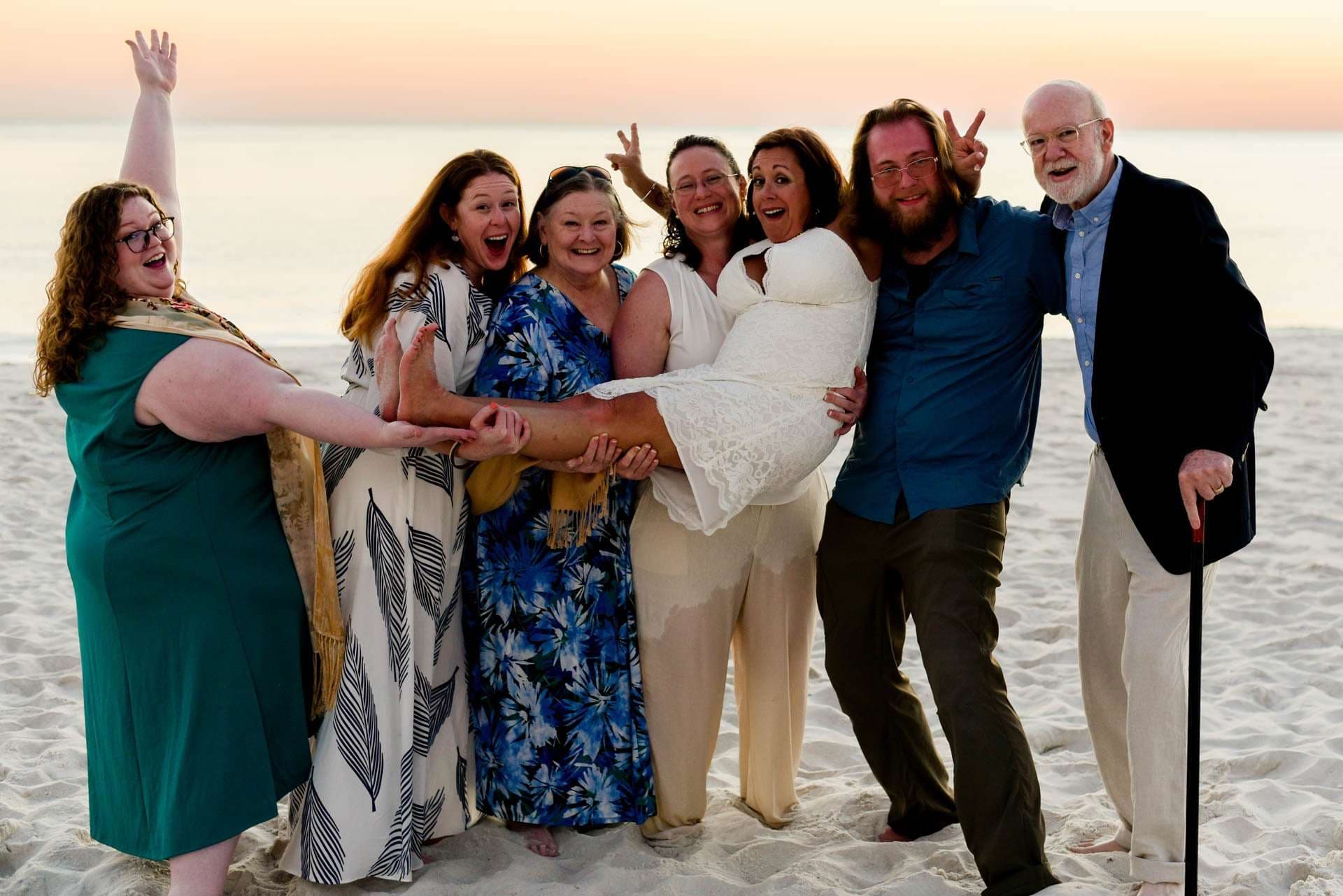 Smiling wedding guests on the beach at sunset cheer as a bearded man cradles a woman in a white lace dress