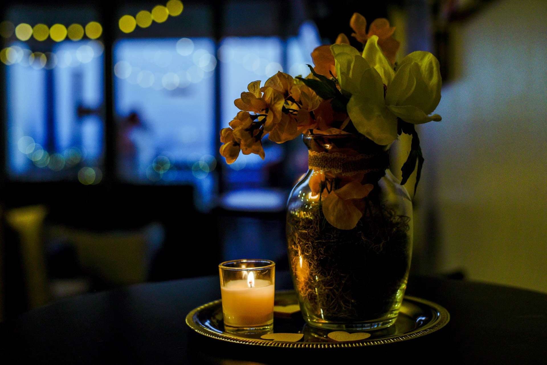 Lit candle and a glass vase of orange and yellow flowers on a silver tray, with softly glowing string lights behind