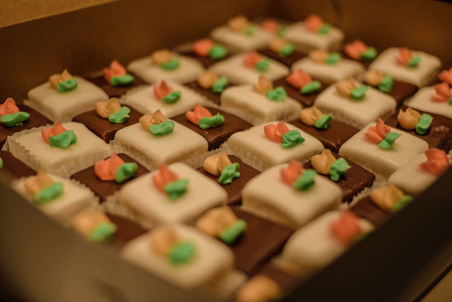 Box of square petit fours with white icing and chocolate bases, topped with small orange and green icing flowers