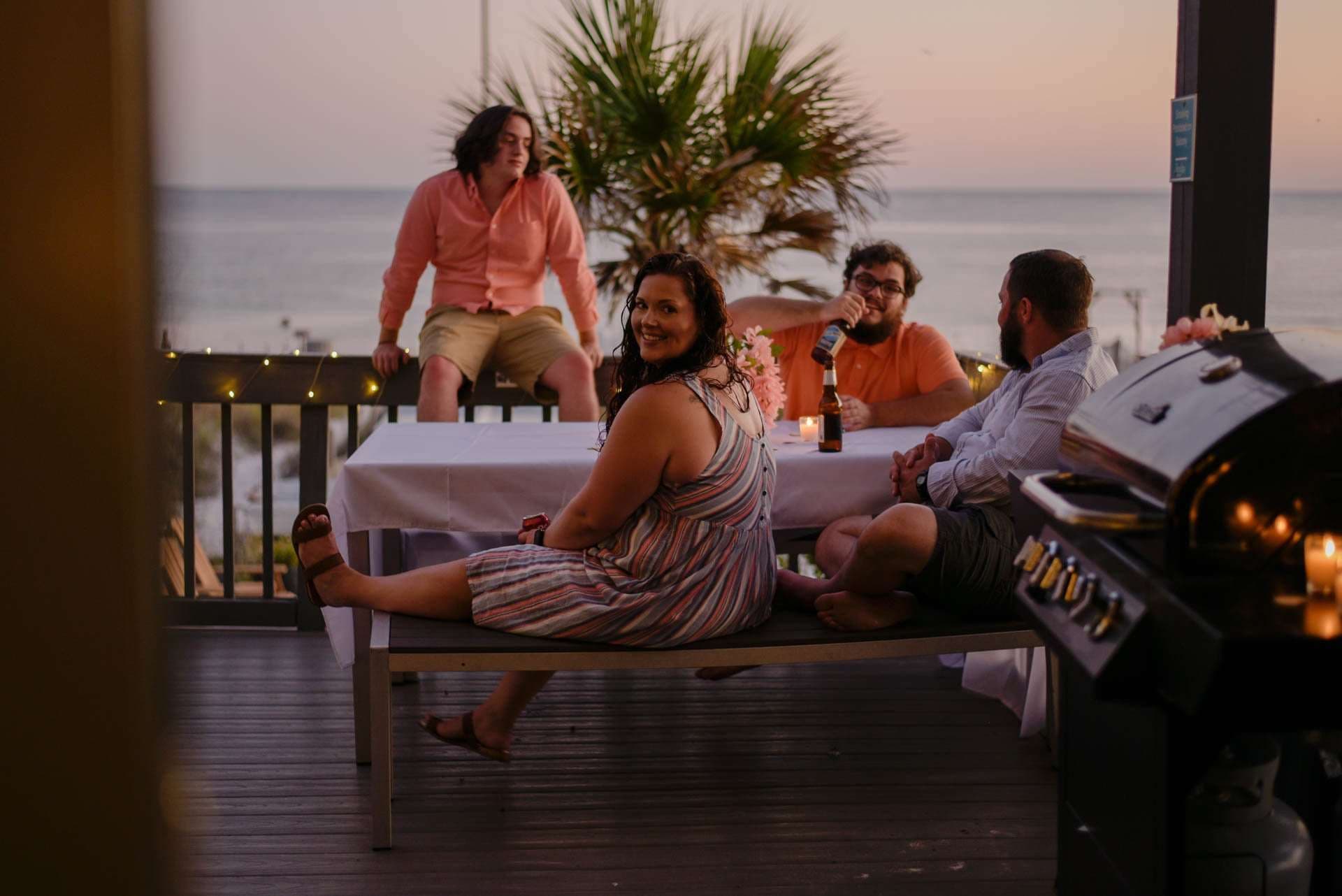 Smiling woman in a striped dress turns to the camera as friends gather around a candlelit table on a deck at sunset