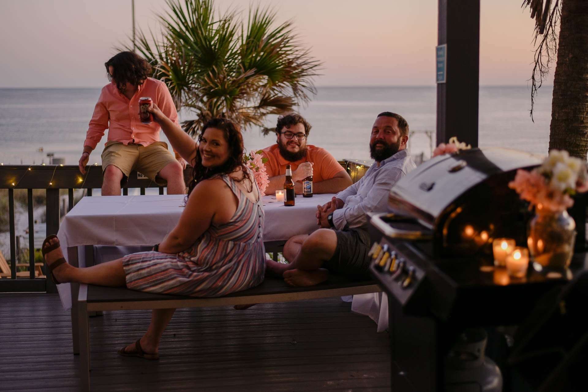 Friends laughing around a candlelit table on a seaside deck at sunset, with a grill and flowers in the foreground