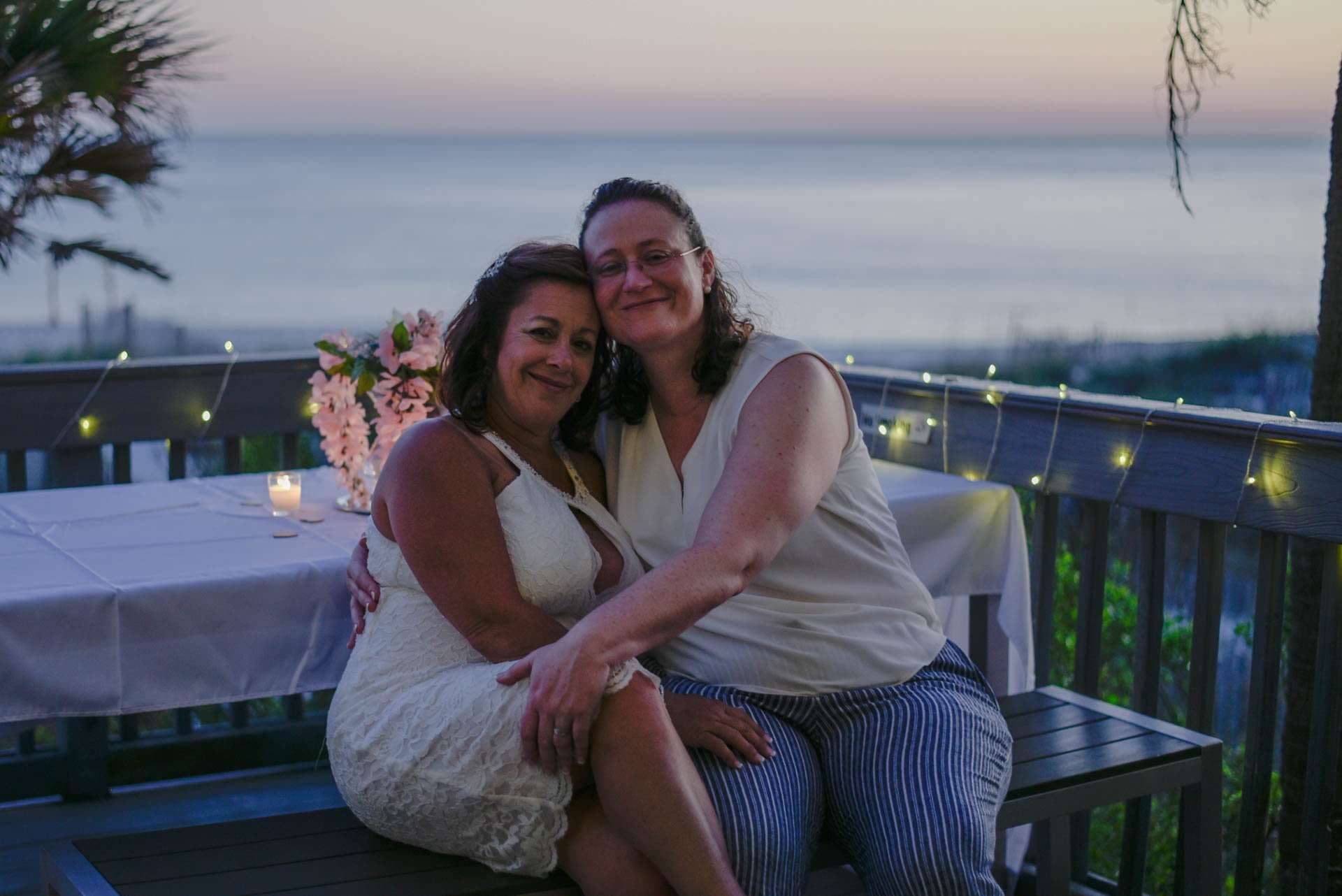 Two women embracing and smiling on a deck with string lights, flowers and candle, ocean at dusk behind them