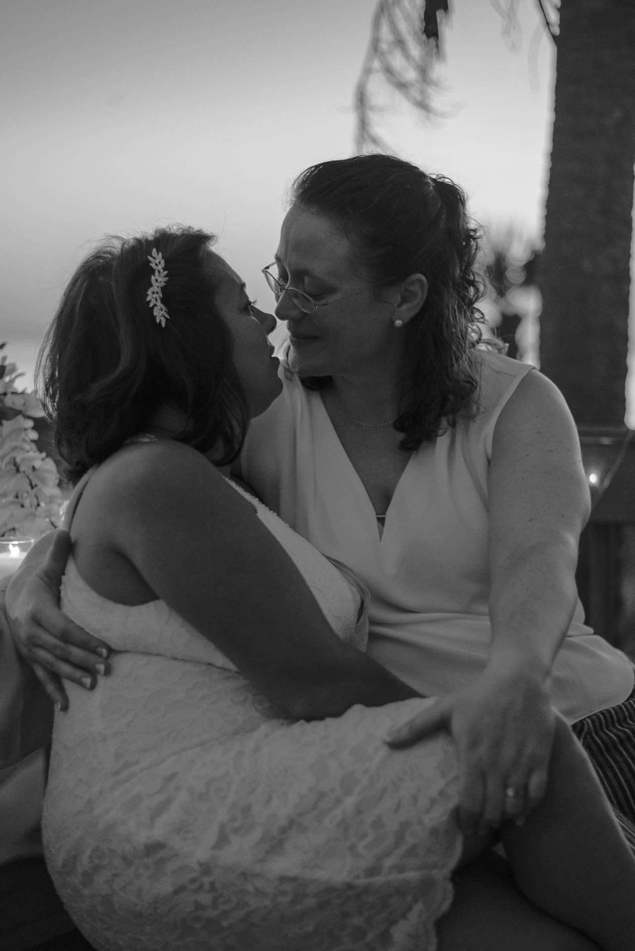 Black-and-white photo of two brides nose to nose in an embrace at dusk, one in a lace dress with a hair clip