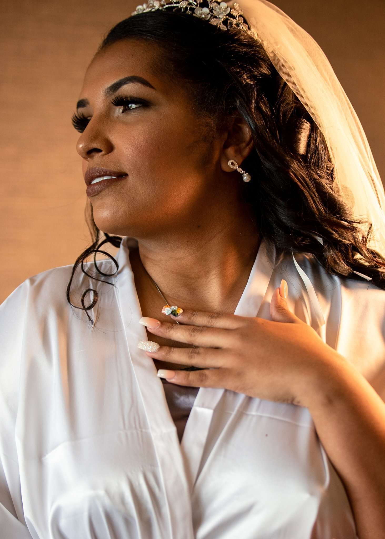 Bride in a white satin robe, veil and tiara glancing aside with her hand resting on her chest