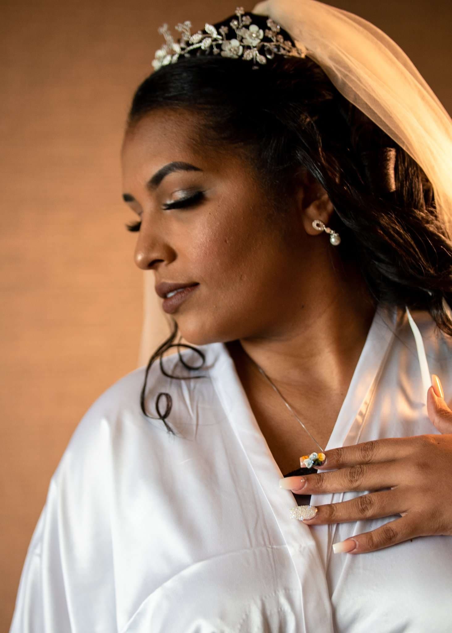 Bride in a white satin robe with a veil and jeweled tiara, eyes closed, looking down with her hand at her collar