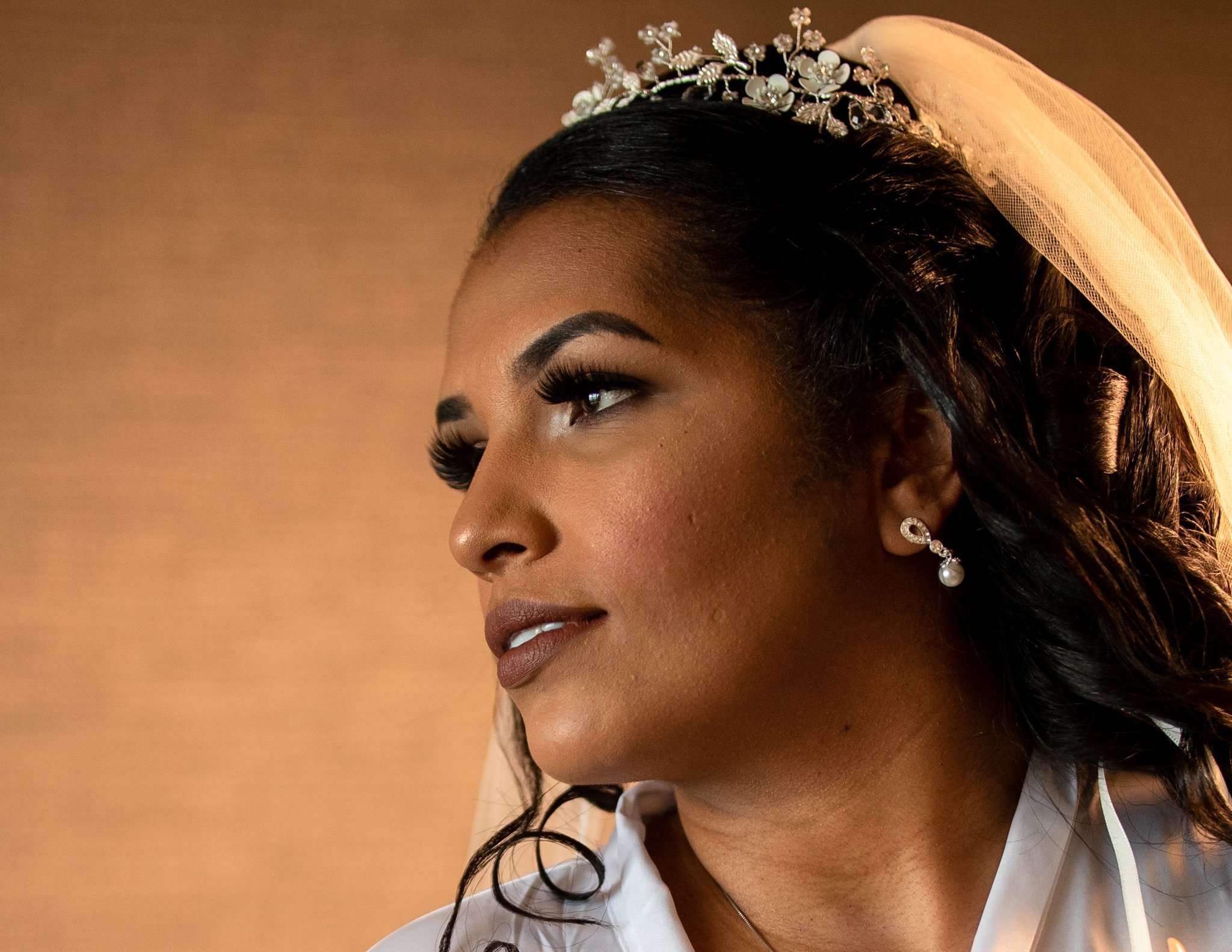 Bride in profile wearing a floral tiara, veil, pearl earring and white robe, gazing off against a warm tan wall