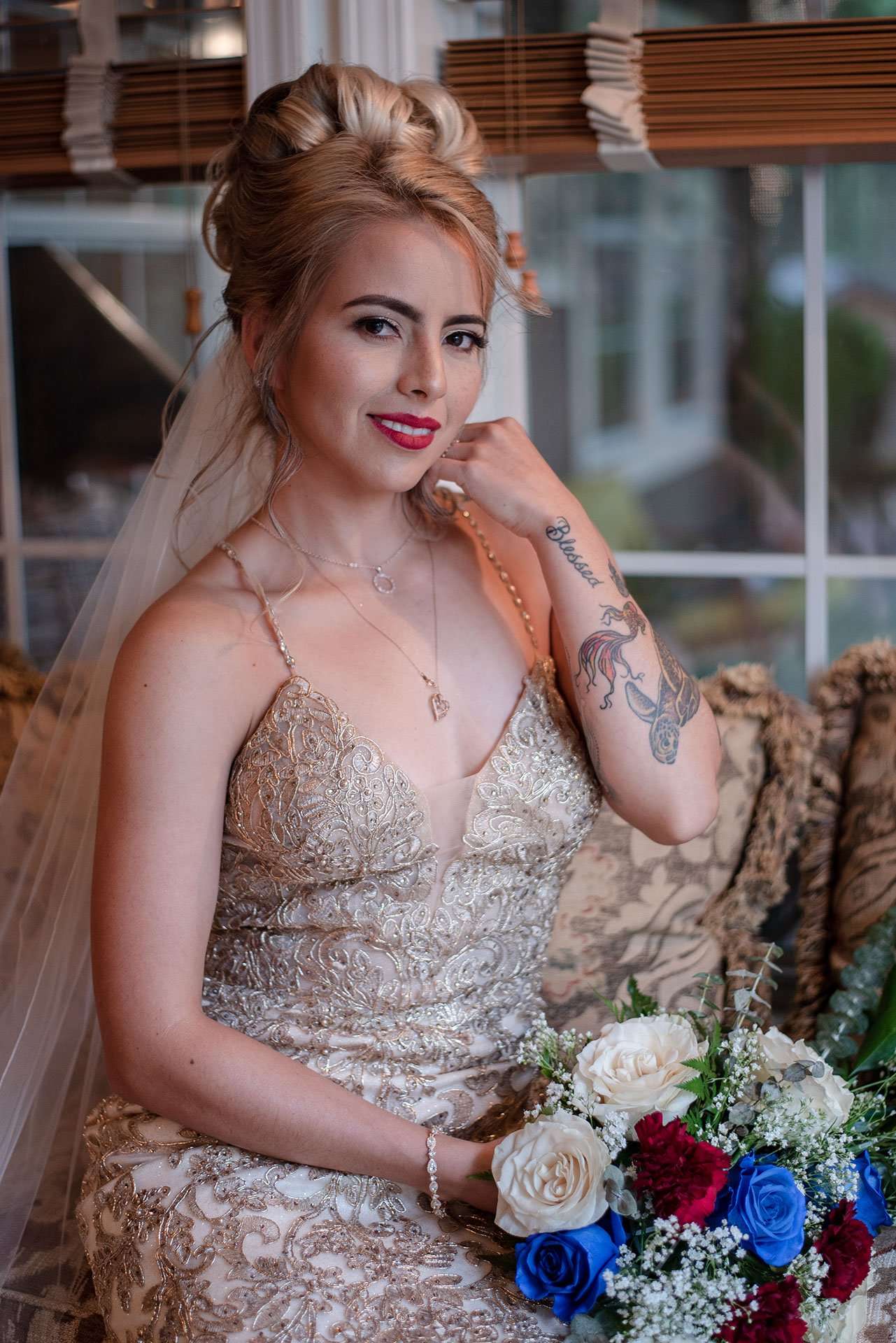 Bride in a gold lace gown and veil holding a bouquet of white, red and blue roses while seated by a window