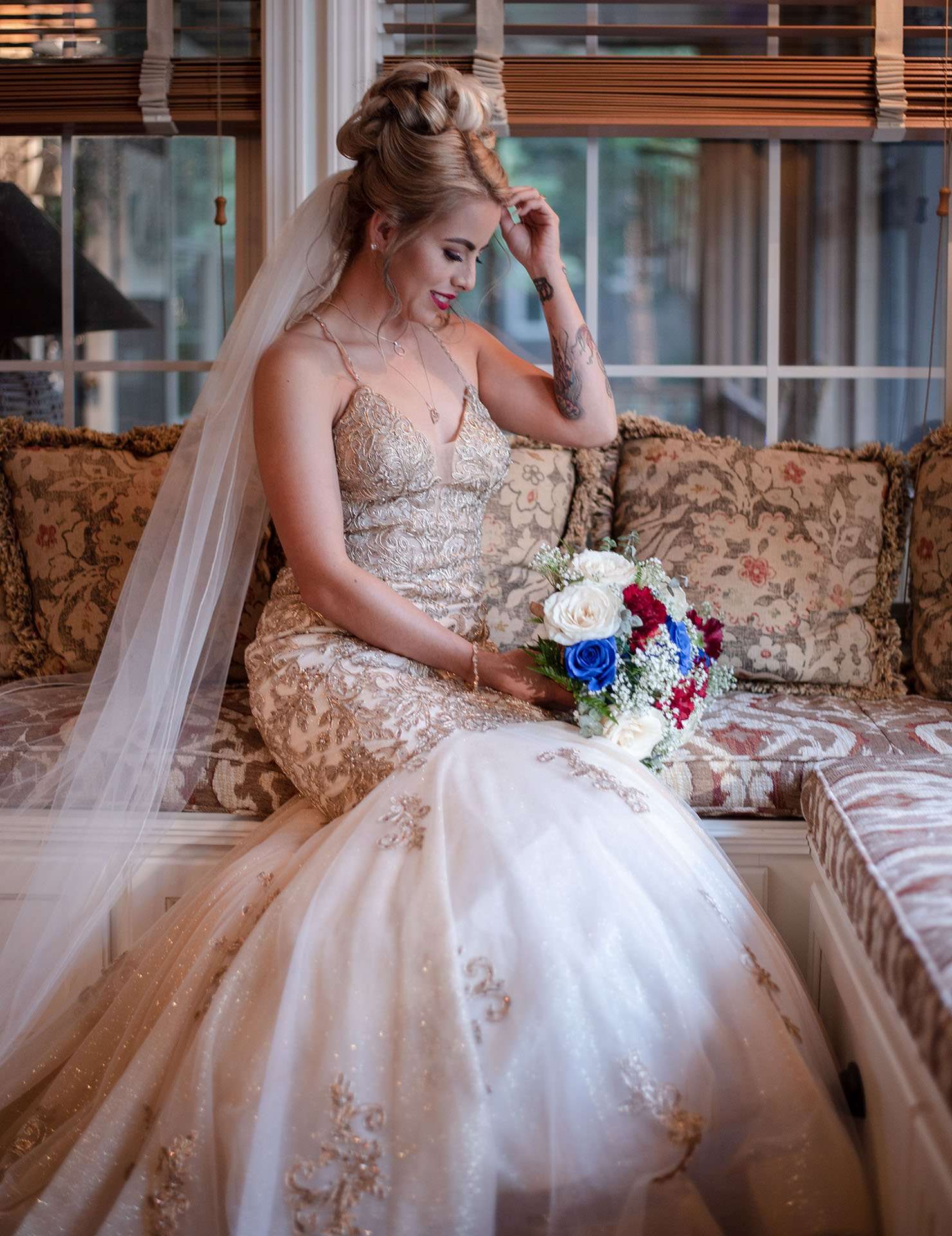 Bride in a gold-embroidered gown and long veil sits on a cushioned window seat holding a red, white and blue bouquet