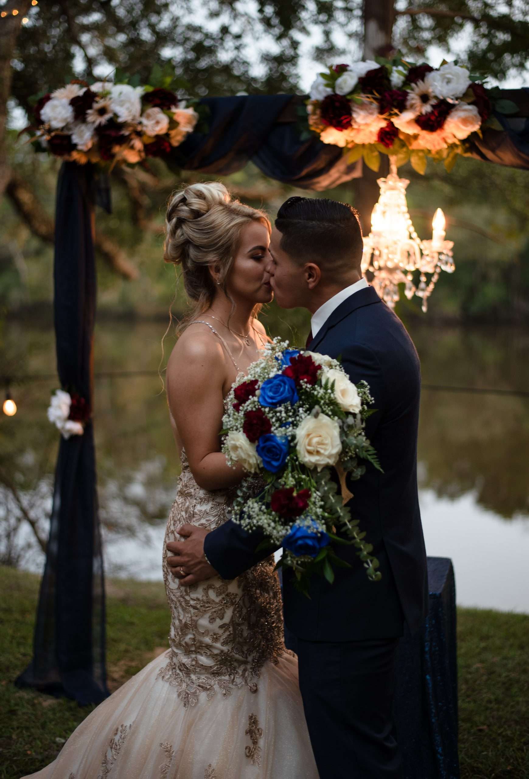 Couple kissing under a draped floral arch with a chandelier by a lake at dusk, bride holding a blue and burgundy bouquet