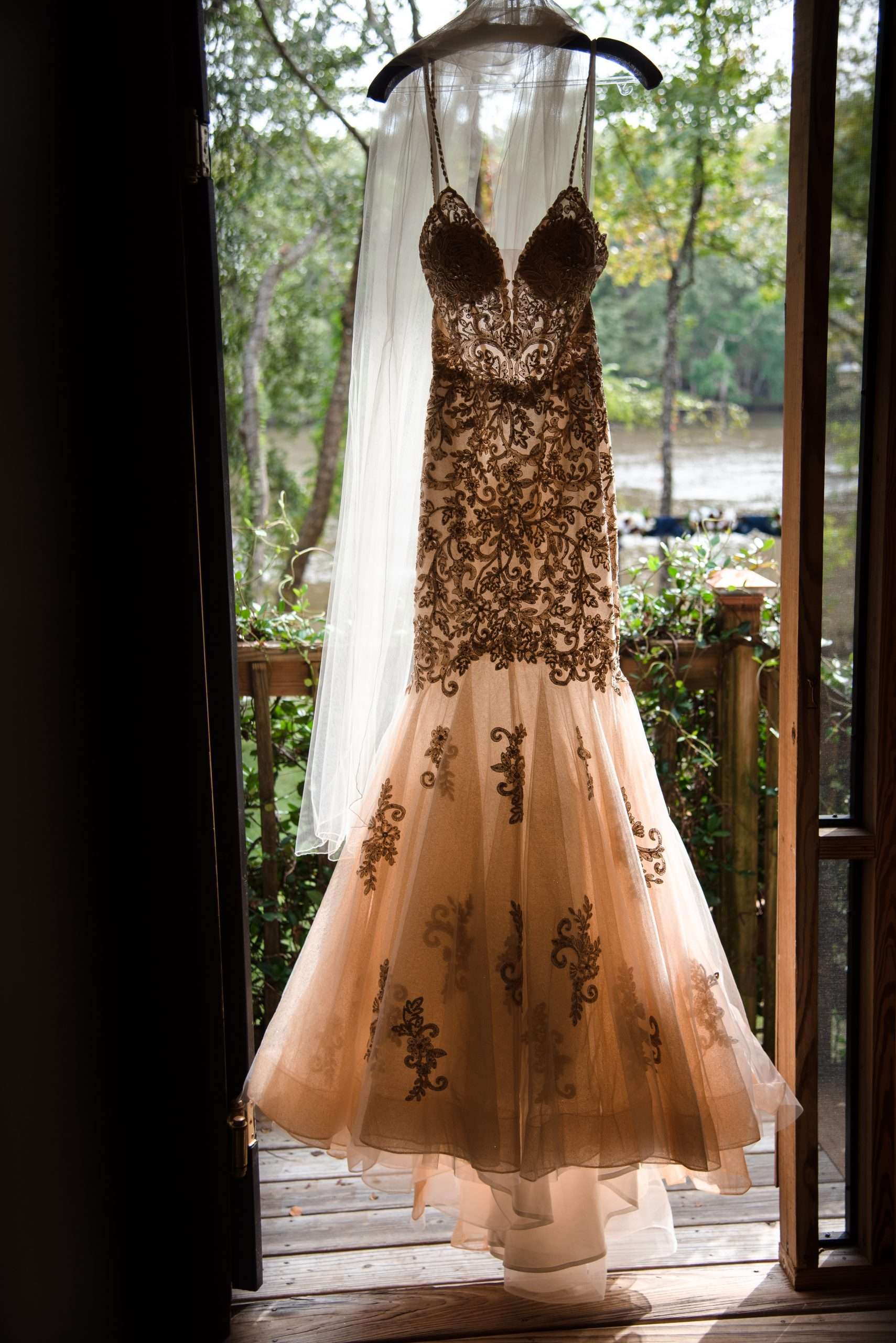 Wedding gown with embroidered bodice and blush tulle skirt hanging in an open doorway above a wooden deck by trees