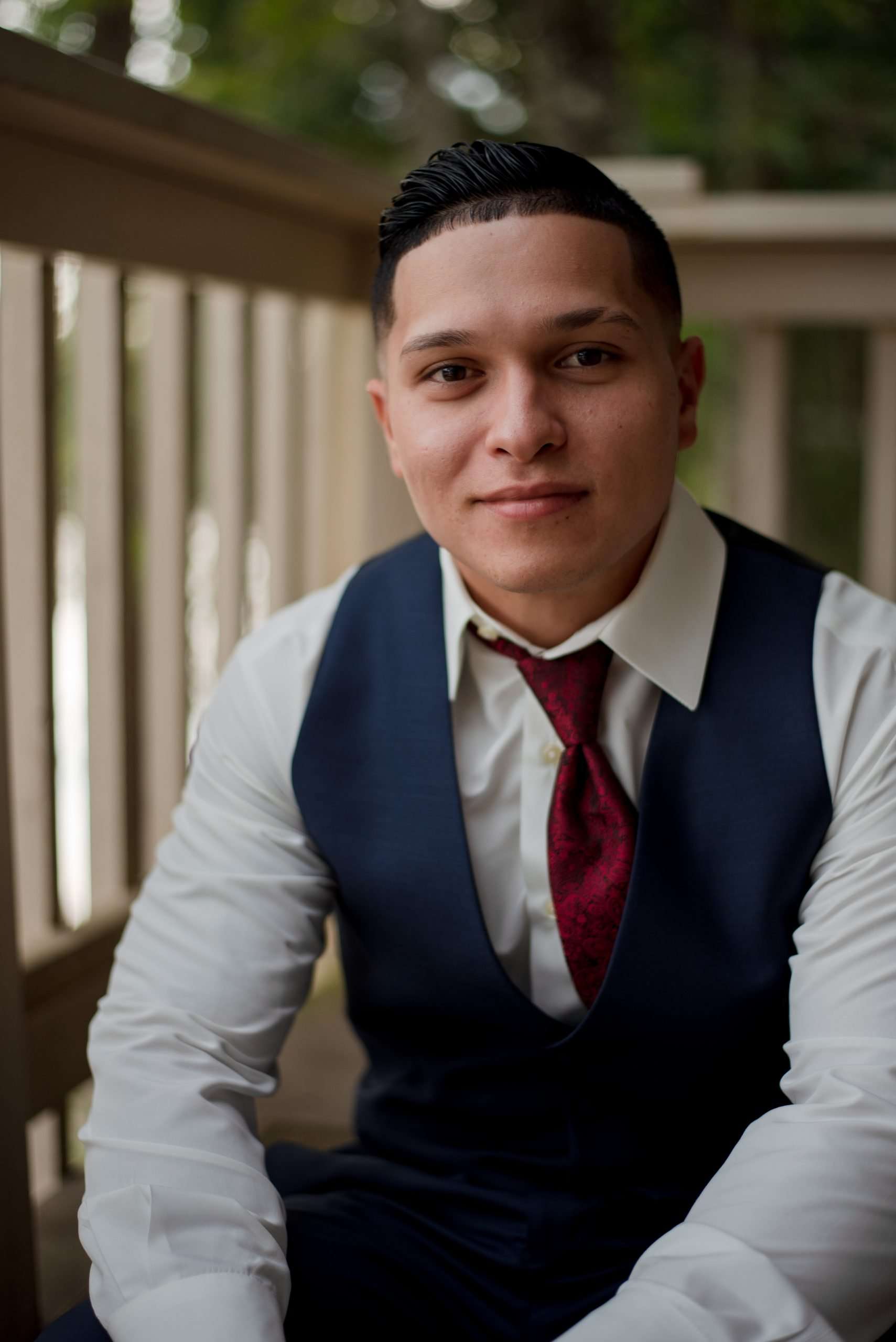 Smiling man in a navy vest, white shirt and burgundy tie seated on a wooden porch with railings behind him