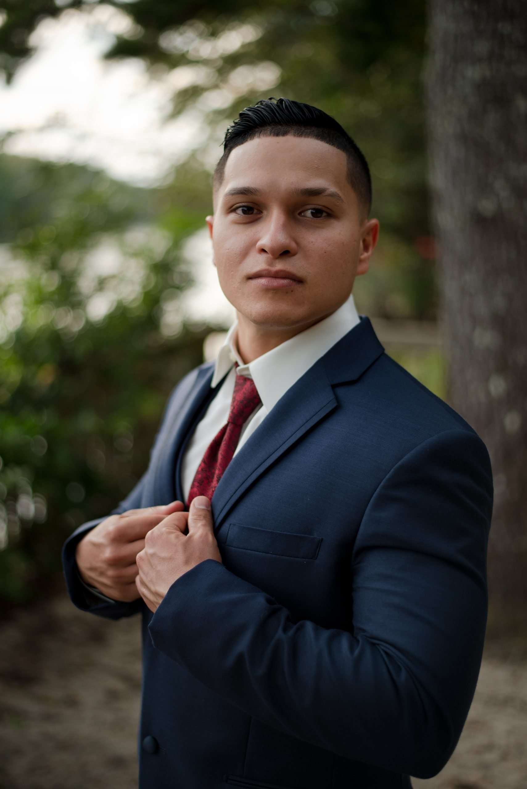 Man in a navy suit and burgundy tie adjusting his jacket lapels outdoors among trees, looking at the camera