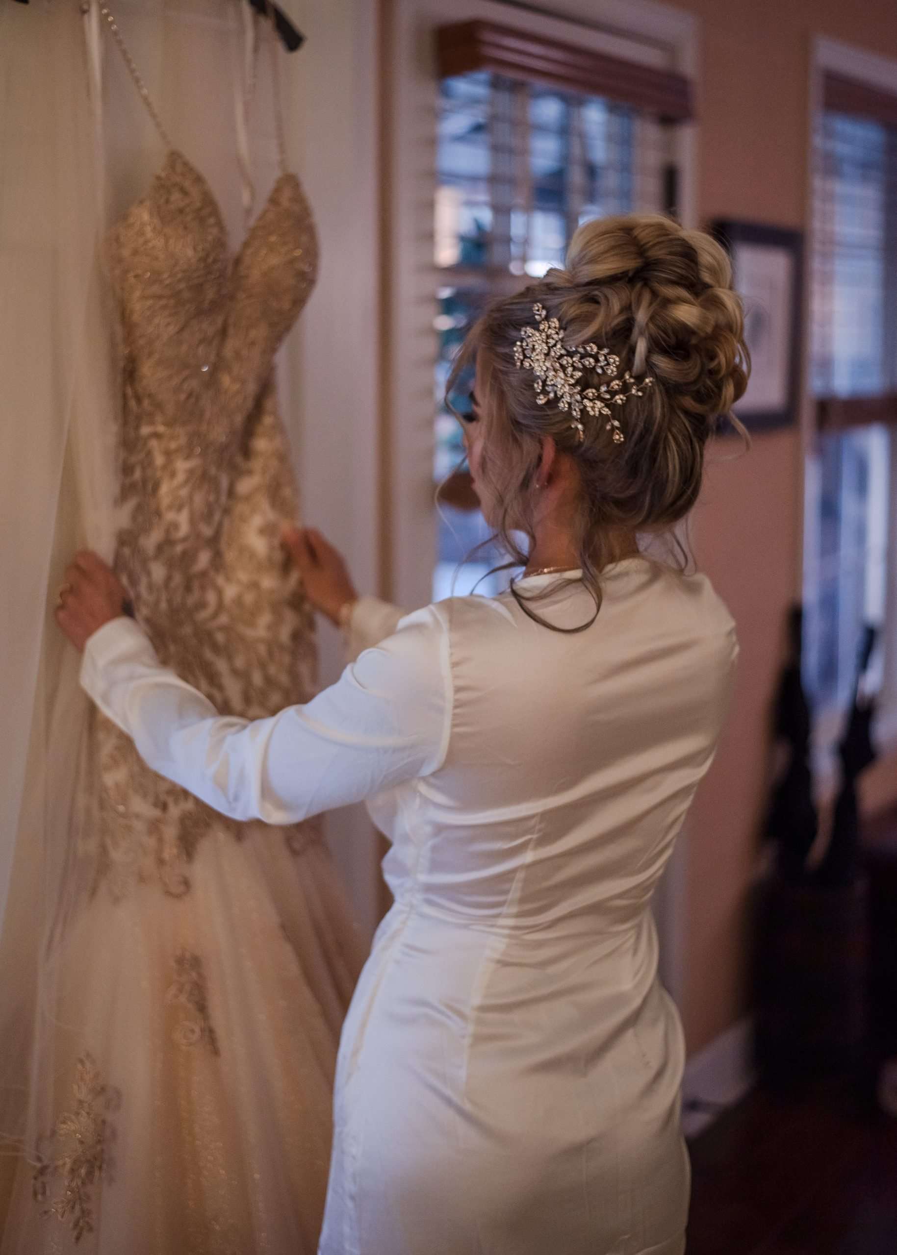 Bride in a white satin robe and jeweled hair piece touches her gold lace wedding gown hanging by a window