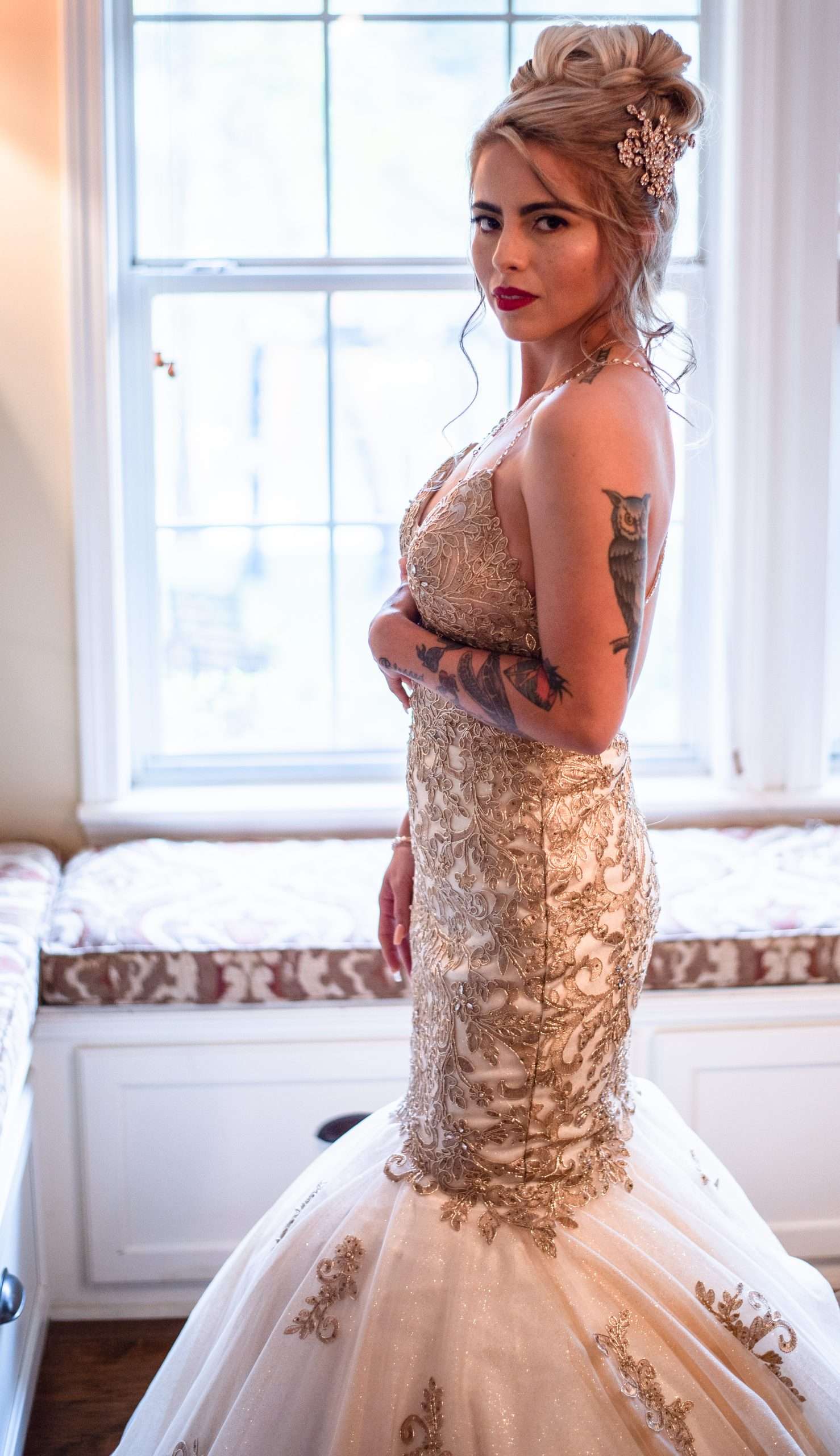 Bride in a gold embroidered mermaid gown with an updo and floral hair piece stands beside a sunlit window seat