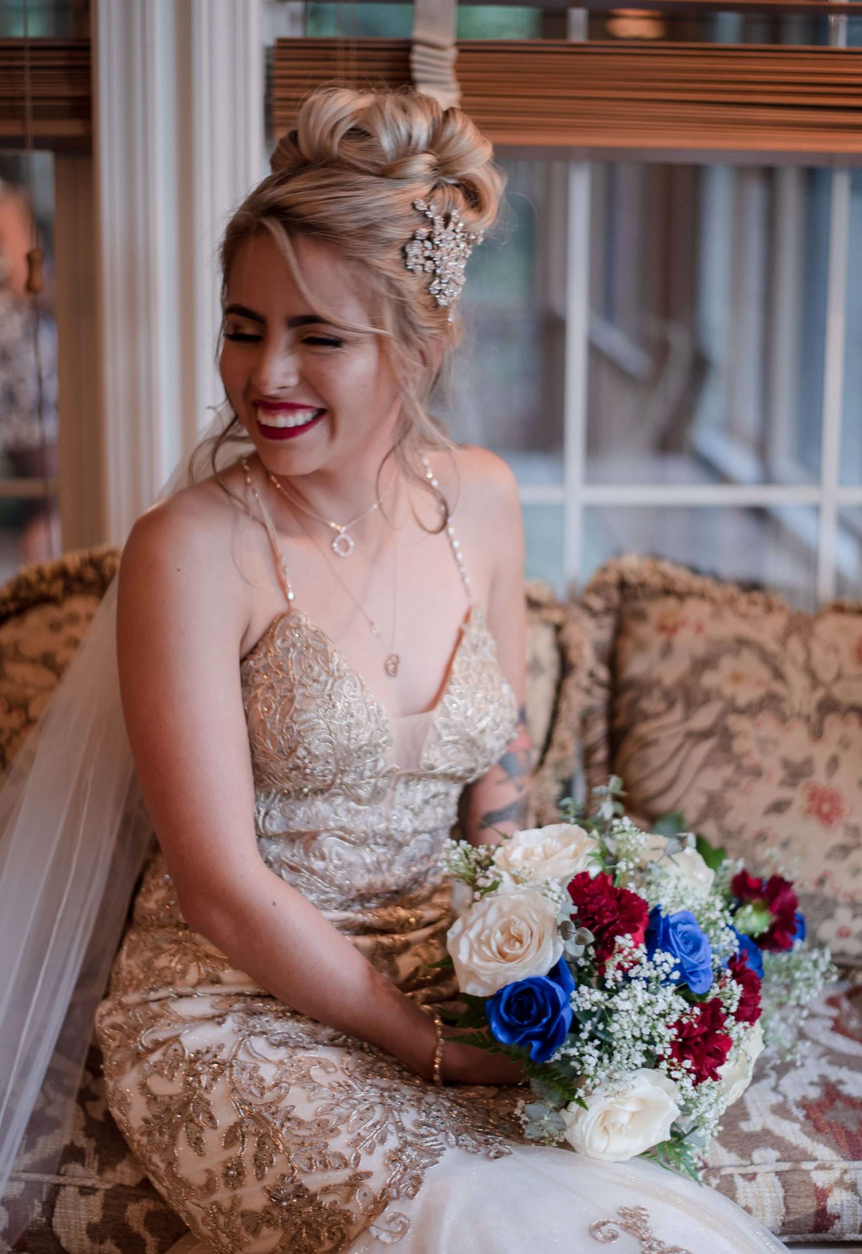 Smiling bride in a gold lace gown with an updo holds a bouquet of white, red and blue roses