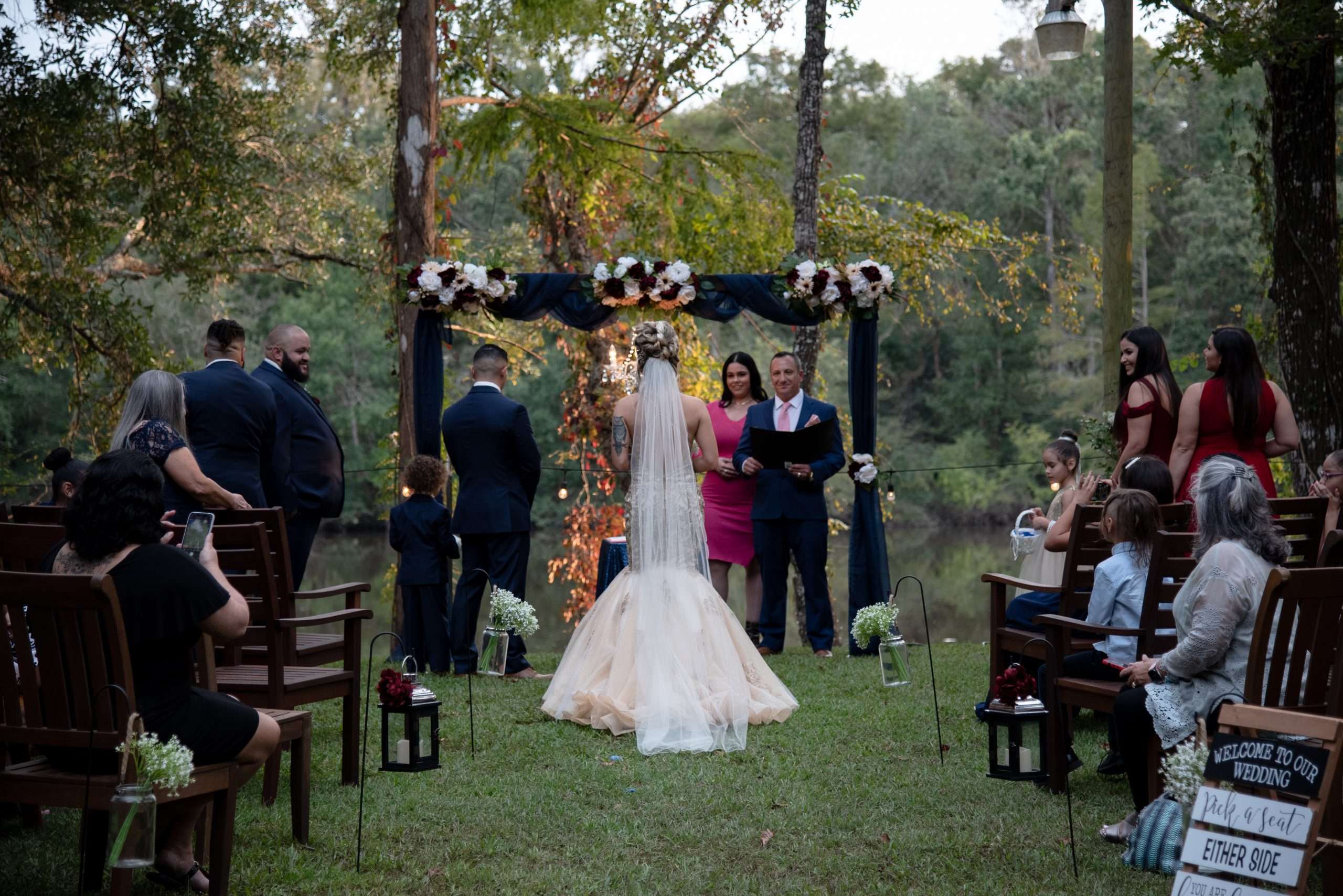 Outdoor wedding ceremony by a river, bride in a long veil facing the officiant beneath a floral arch as guests watch