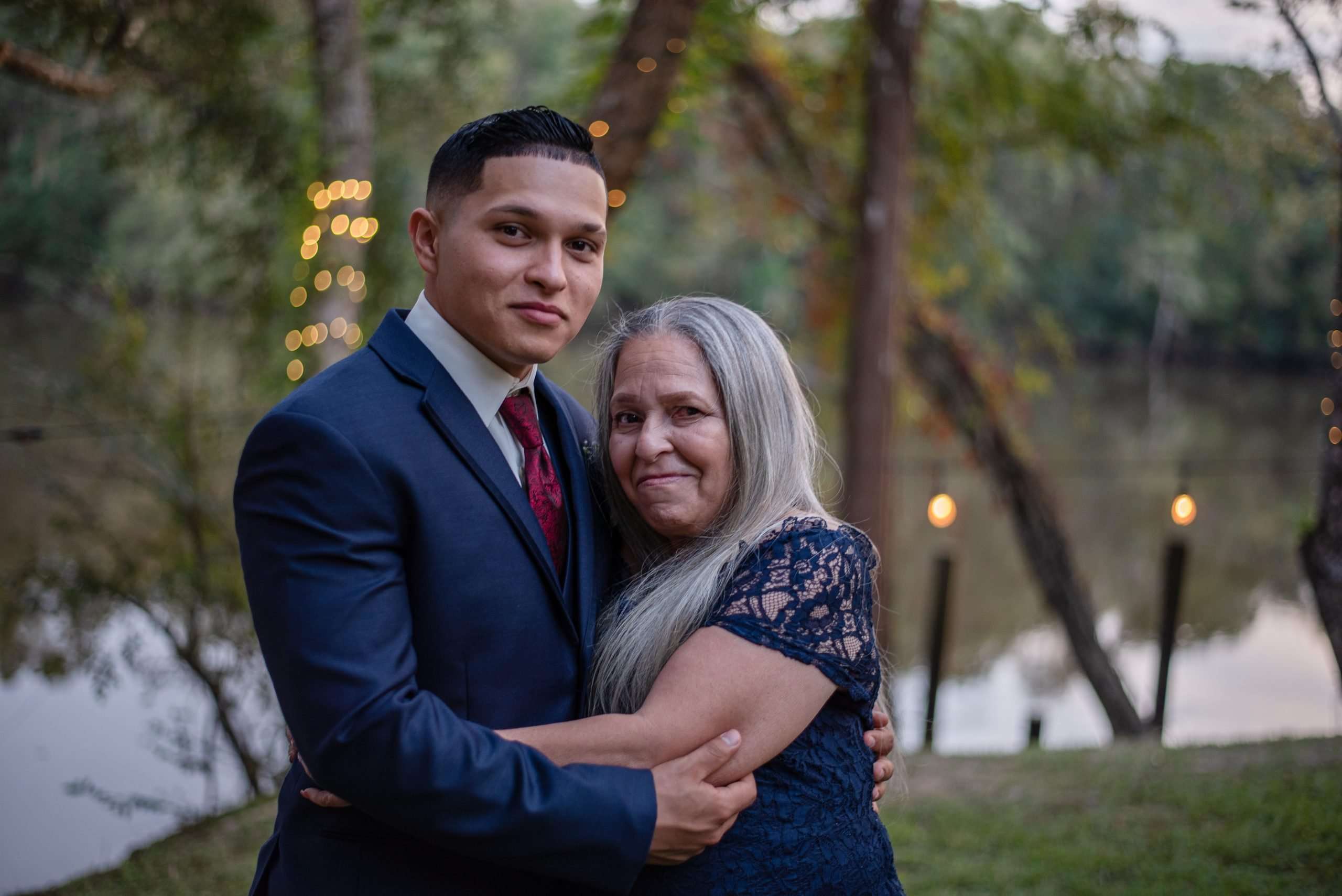 Young man in a navy suit embracing an older woman with long gray hair in a blue lace dress by a lake