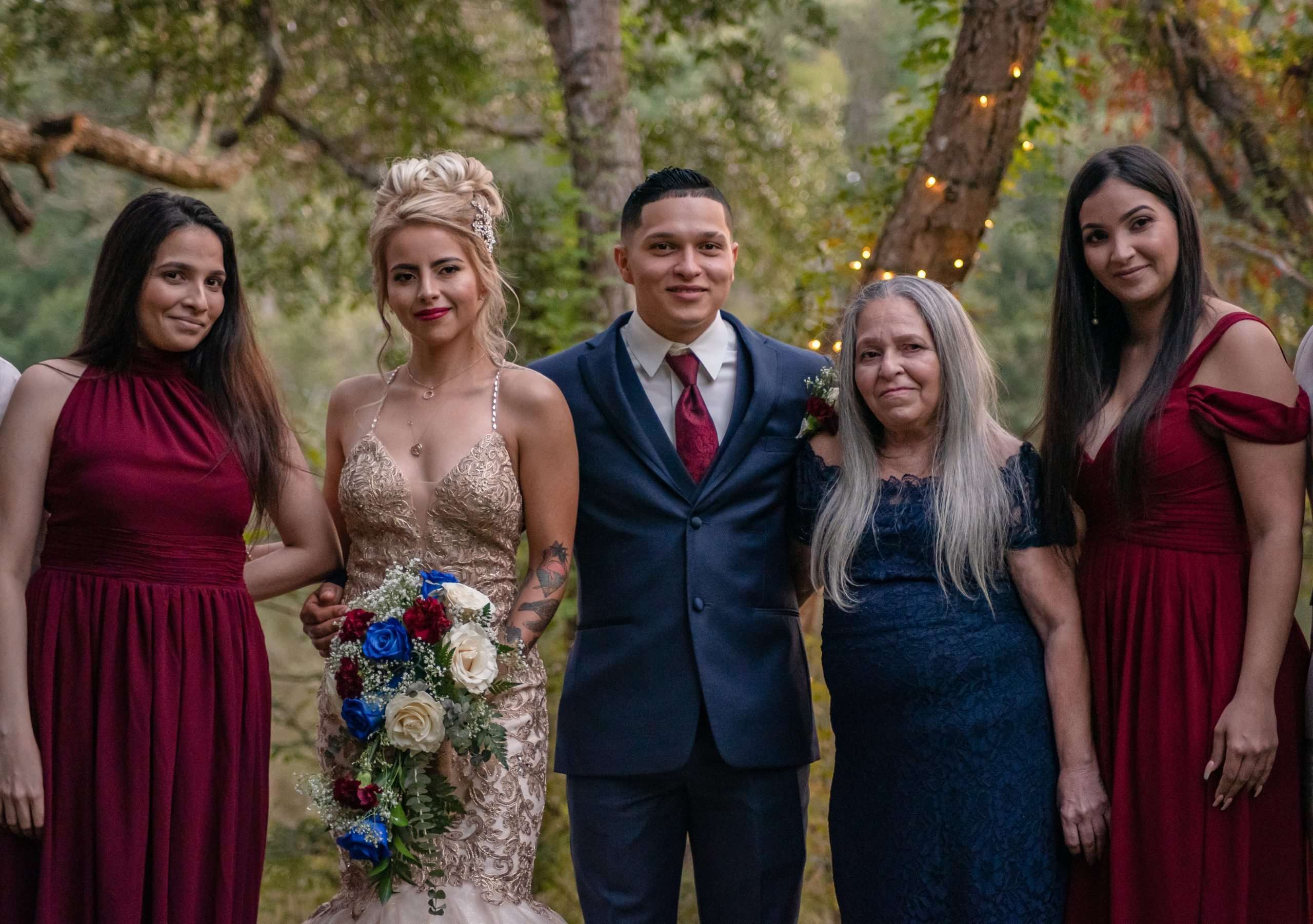 Bride in a gold beaded gown and groom in a navy suit pose with four women in burgundy and navy dresses outdoors