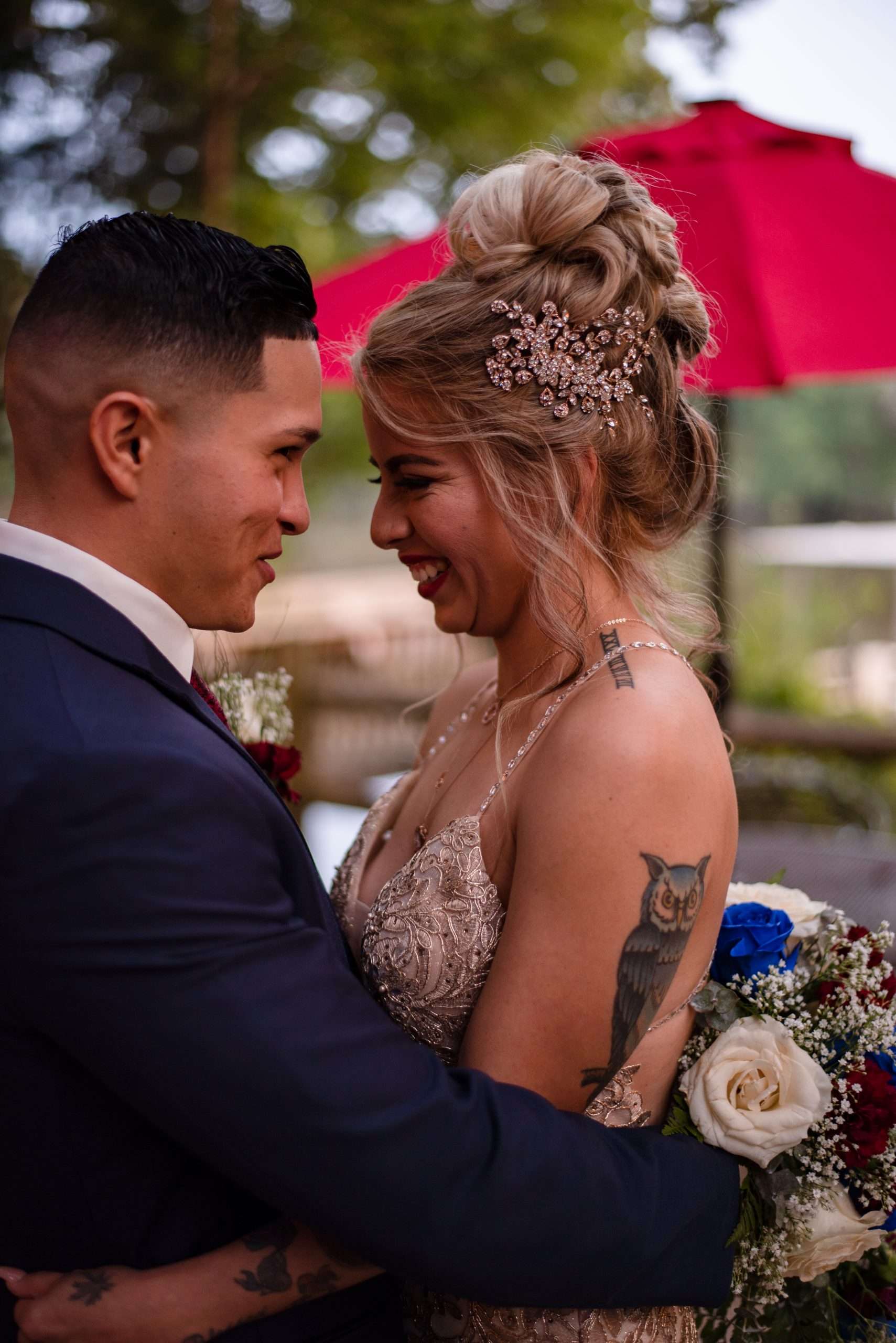 Smiling couple embracing outdoors, bride with an updo, jeweled hairpiece and owl tattoo, holding a rose bouquet