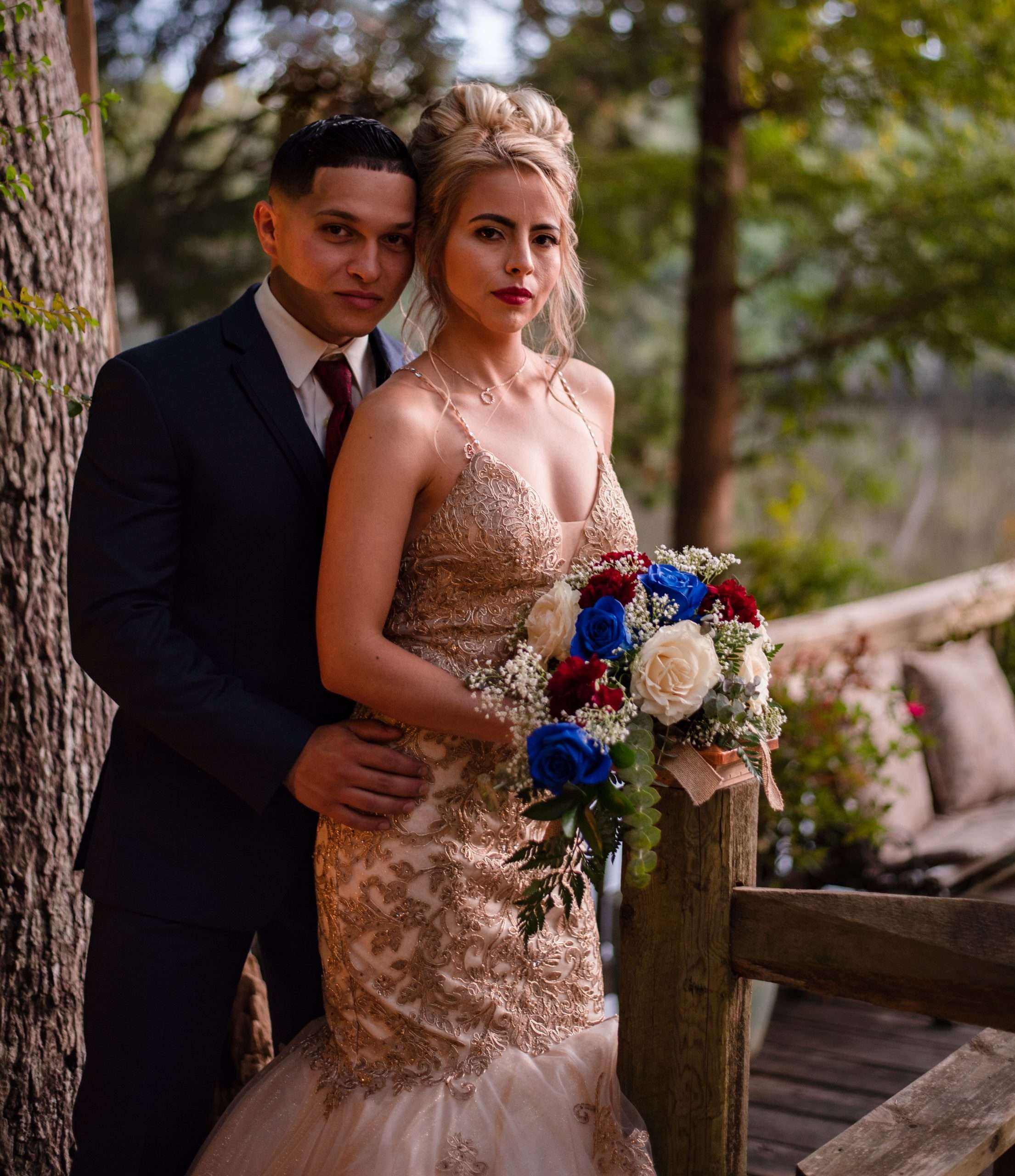 Groom in dark suit embracing bride in gold lace gown holding red, white and blue bouquet beside a wooden railing