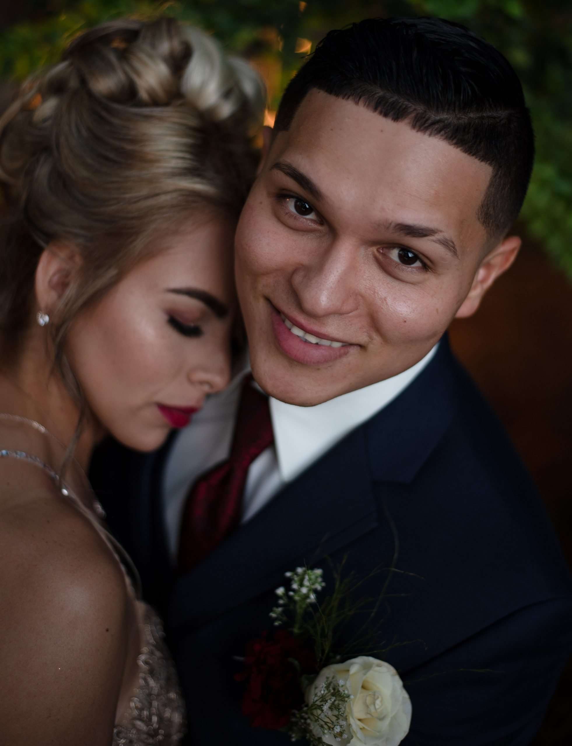 Smiling groom in a navy suit with a burgundy tie and rose boutonniere embraces a woman with closed eyes and red lipstick