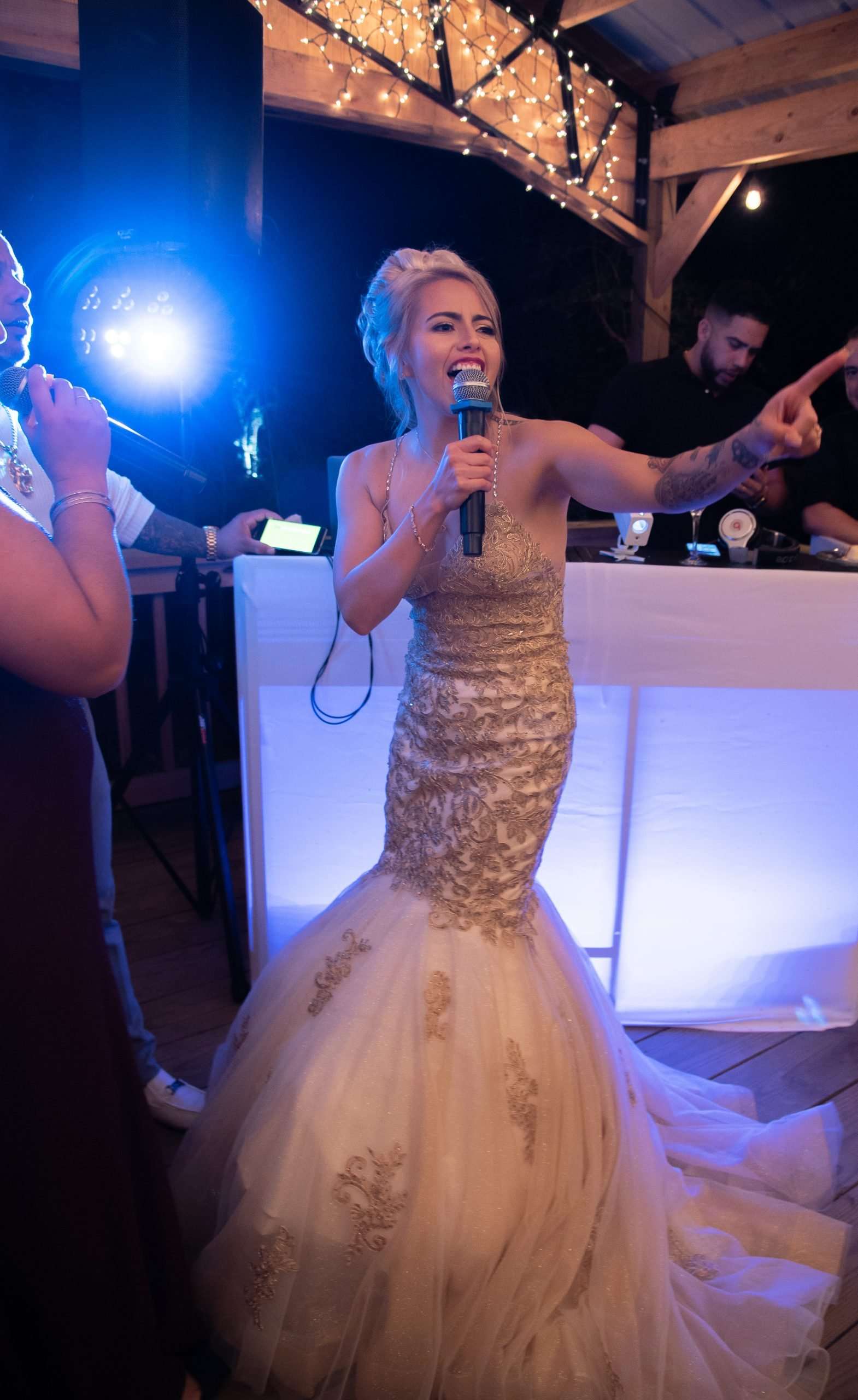 Bride in a gold-embroidered mermaid gown singing into a microphone and pointing at a reception with a DJ behind her
