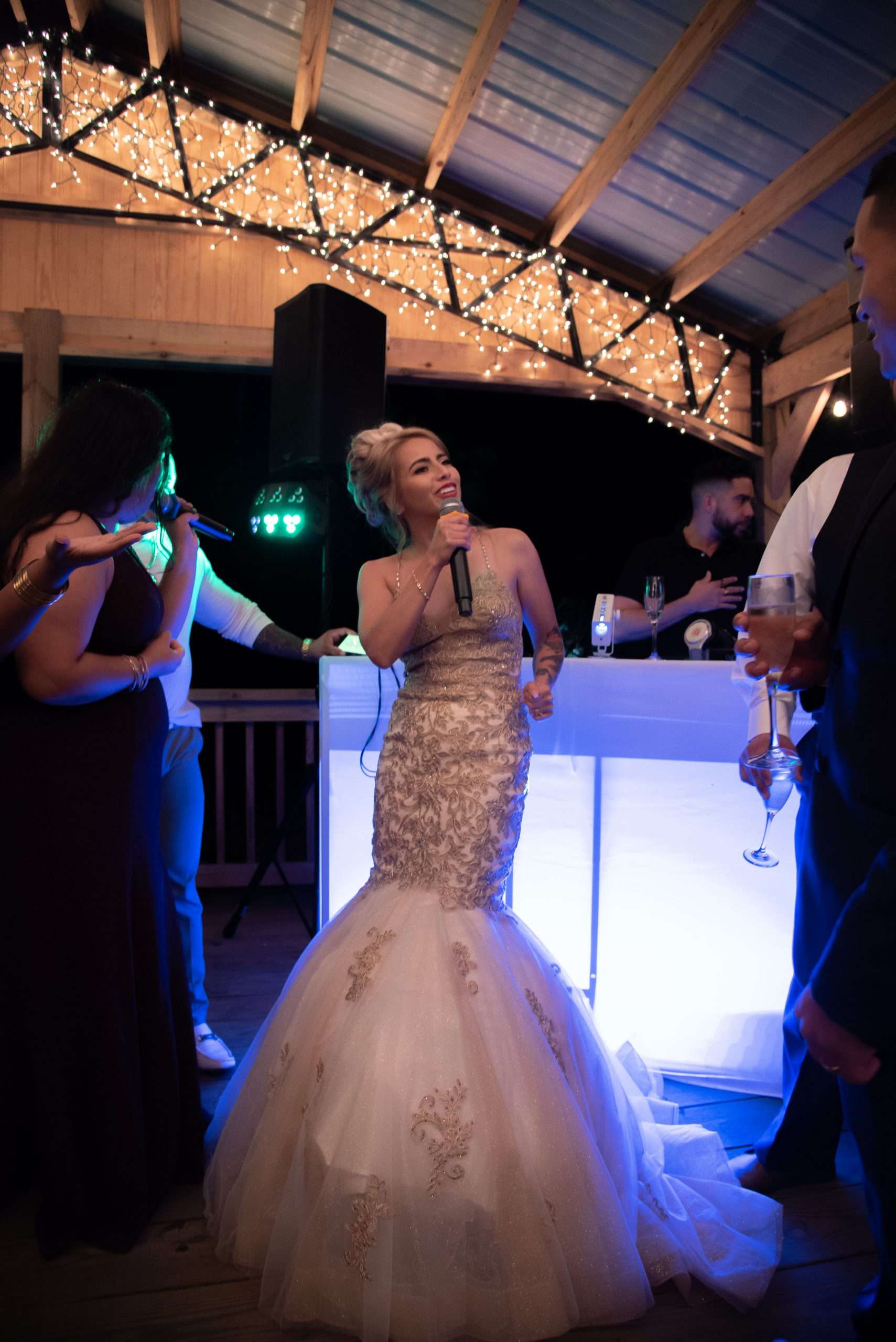 Bride in a gold-embroidered mermaid gown sings into a microphone under string lights at an evening reception