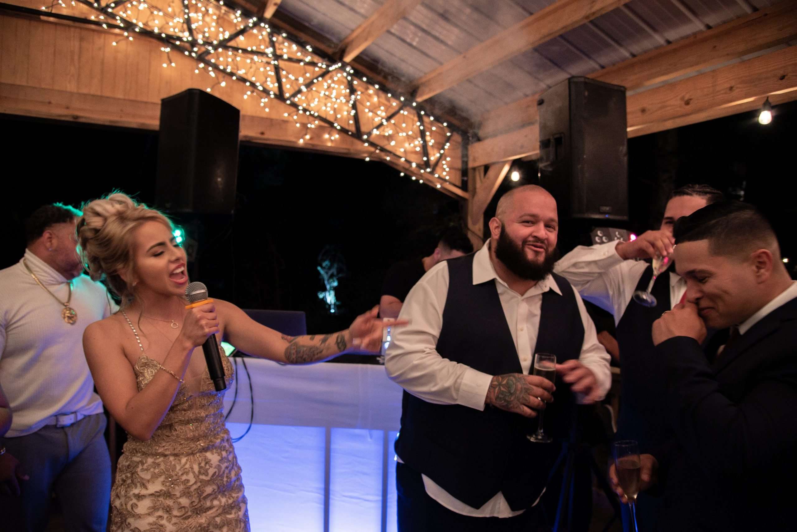 Woman in a gold sequined dress sings into a microphone while smiling guests hold champagne glasses under string lights