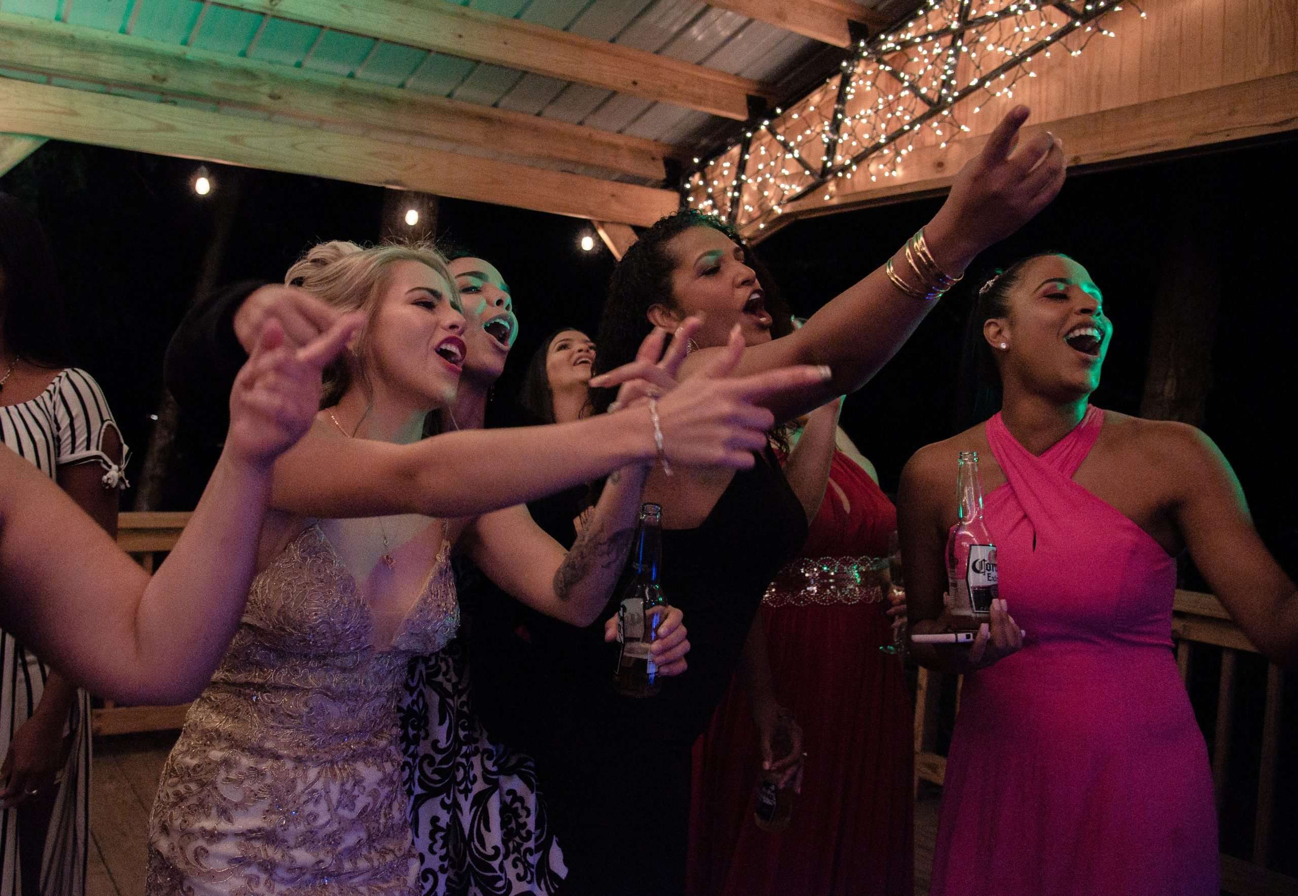 Women in formal gowns pointing and singing joyfully while holding beer bottles under string lights on a covered wooden deck