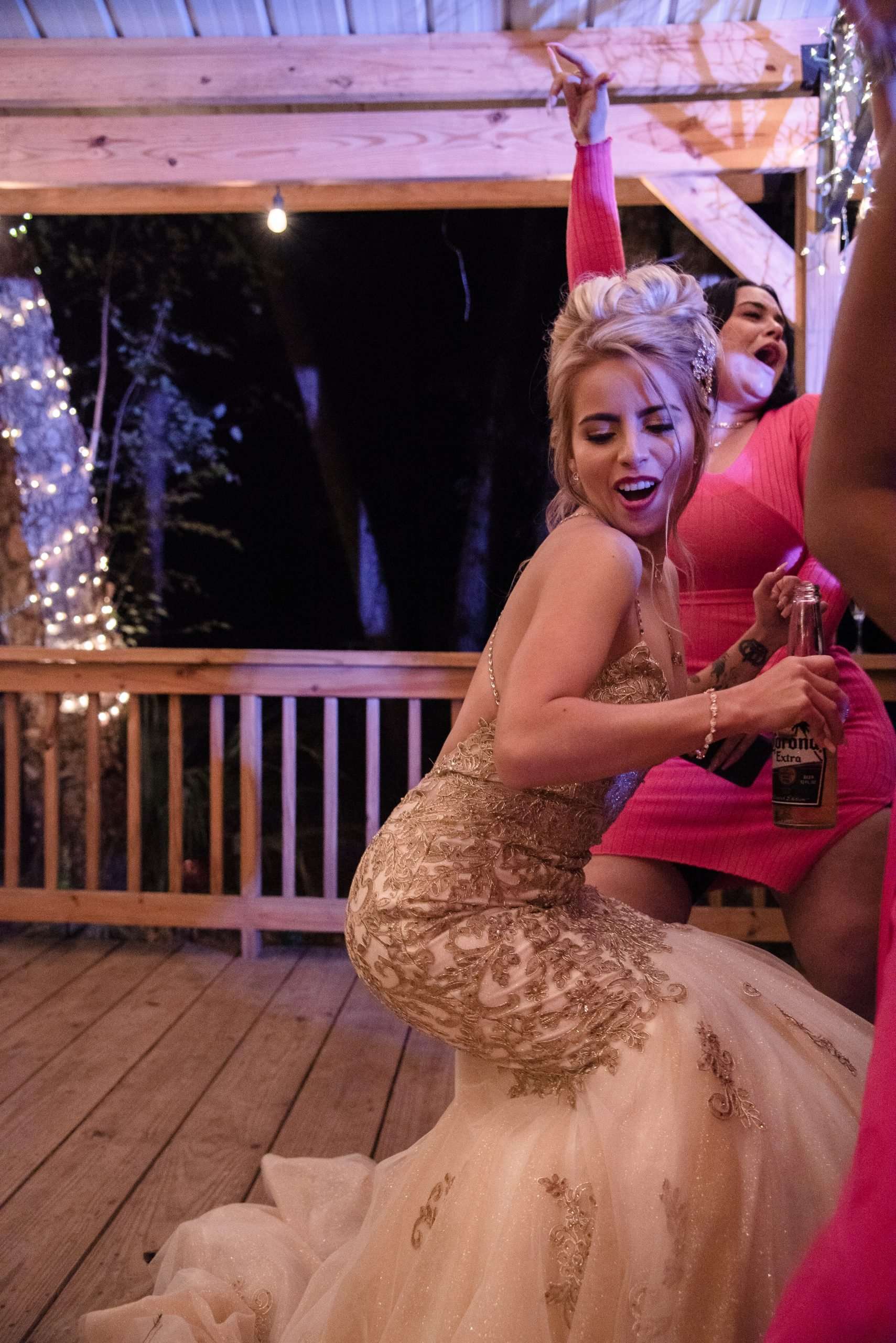 Woman in a gold lace wedding gown dancing with a beer bottle on a lit wooden deck as a guest in pink cheers behind her