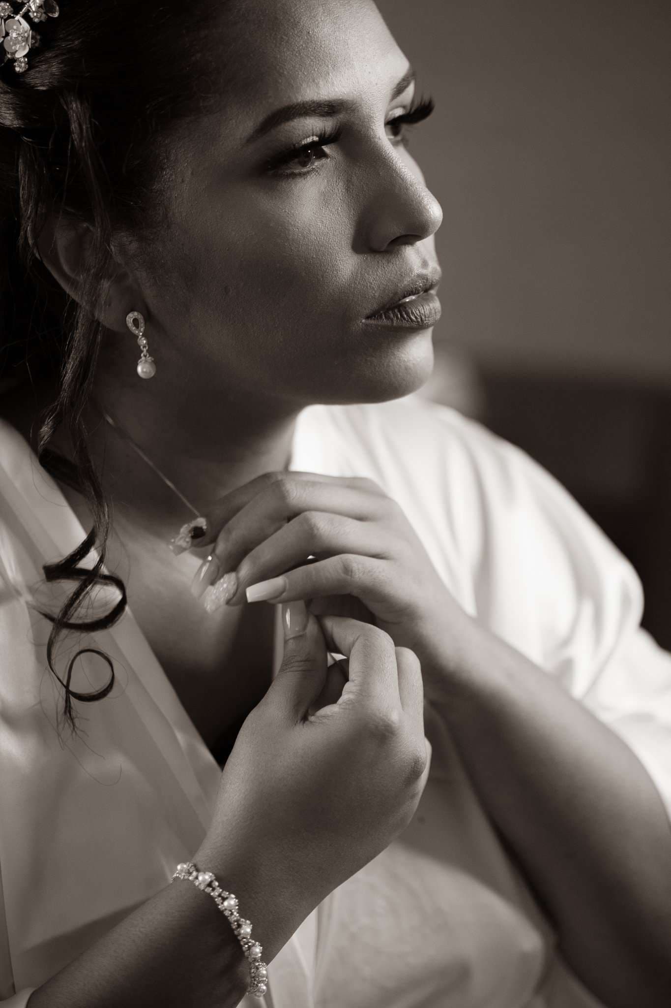 Black-and-white portrait of a bride in a satin robe fastening her necklace, gazing off to the side