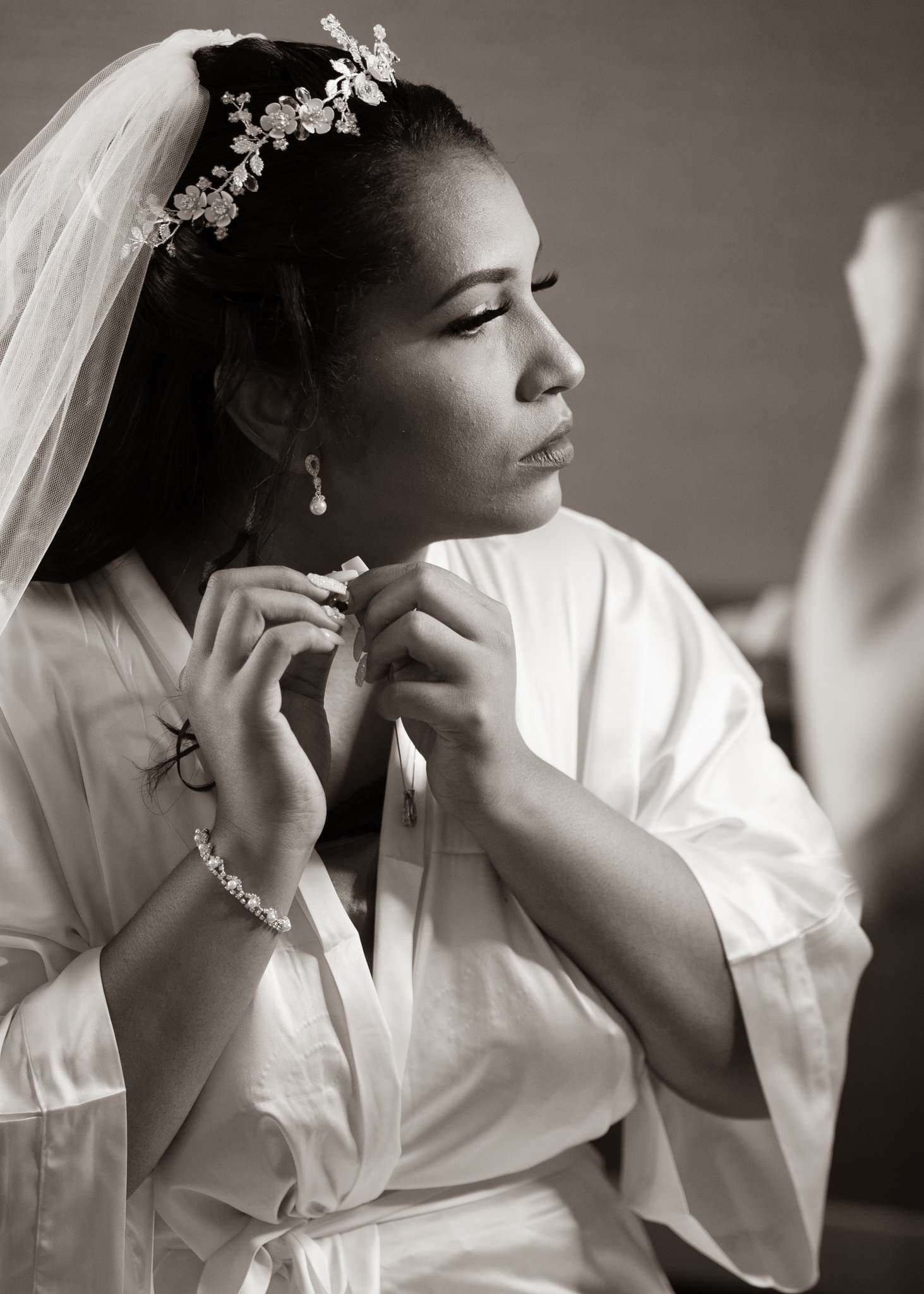 Black-and-white portrait of a bride in a satin robe and veil fastening her earring while getting ready