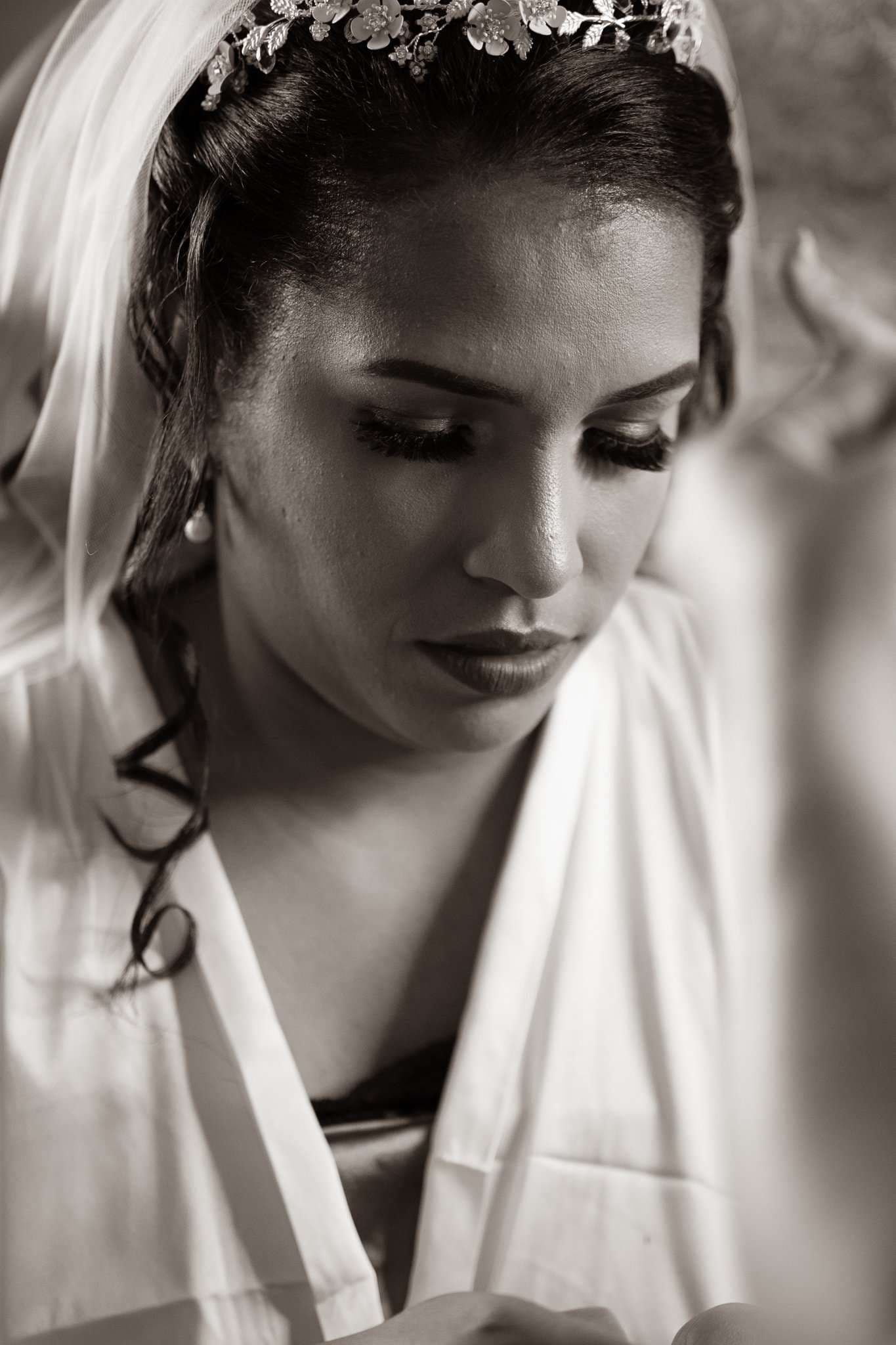 Black-and-white portrait of a bride in a floral hair piece and veil, looking down in a white robe