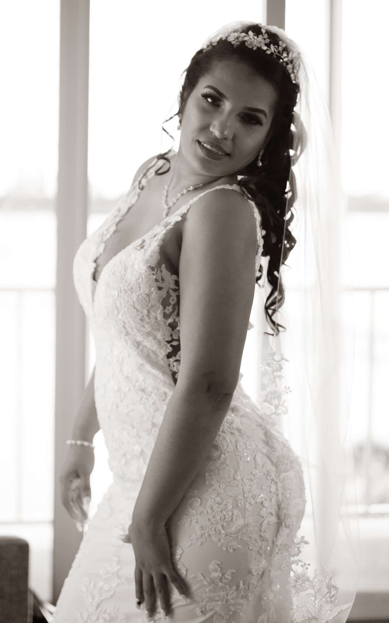 Black-and-white portrait of a bride in a lace gown and floral headpiece with veil, glancing over her shoulder by a window