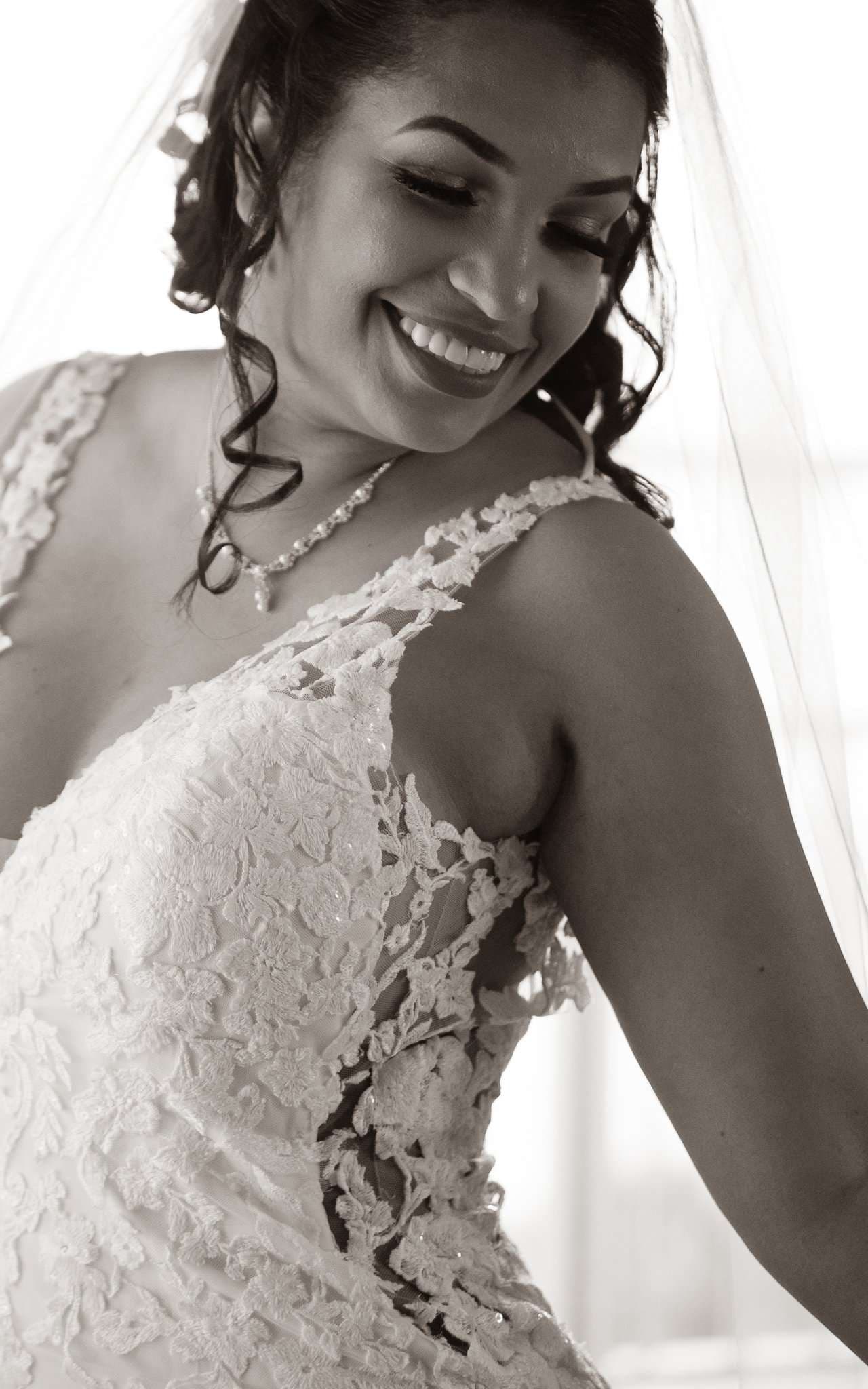 Black-and-white portrait of a smiling bride in a lace gown and veil glancing over her shoulder