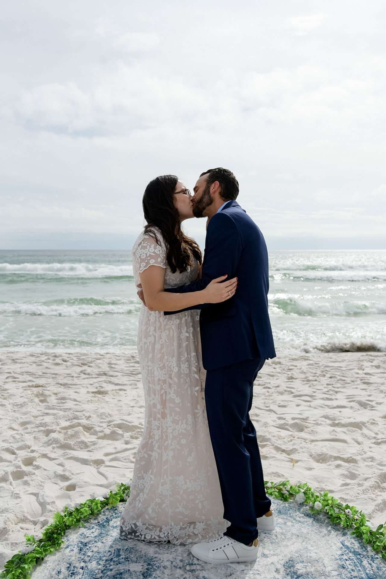Couple kissing on the beach, she in a white lace gown and he in a navy suit, standing on a greenery-edged rug