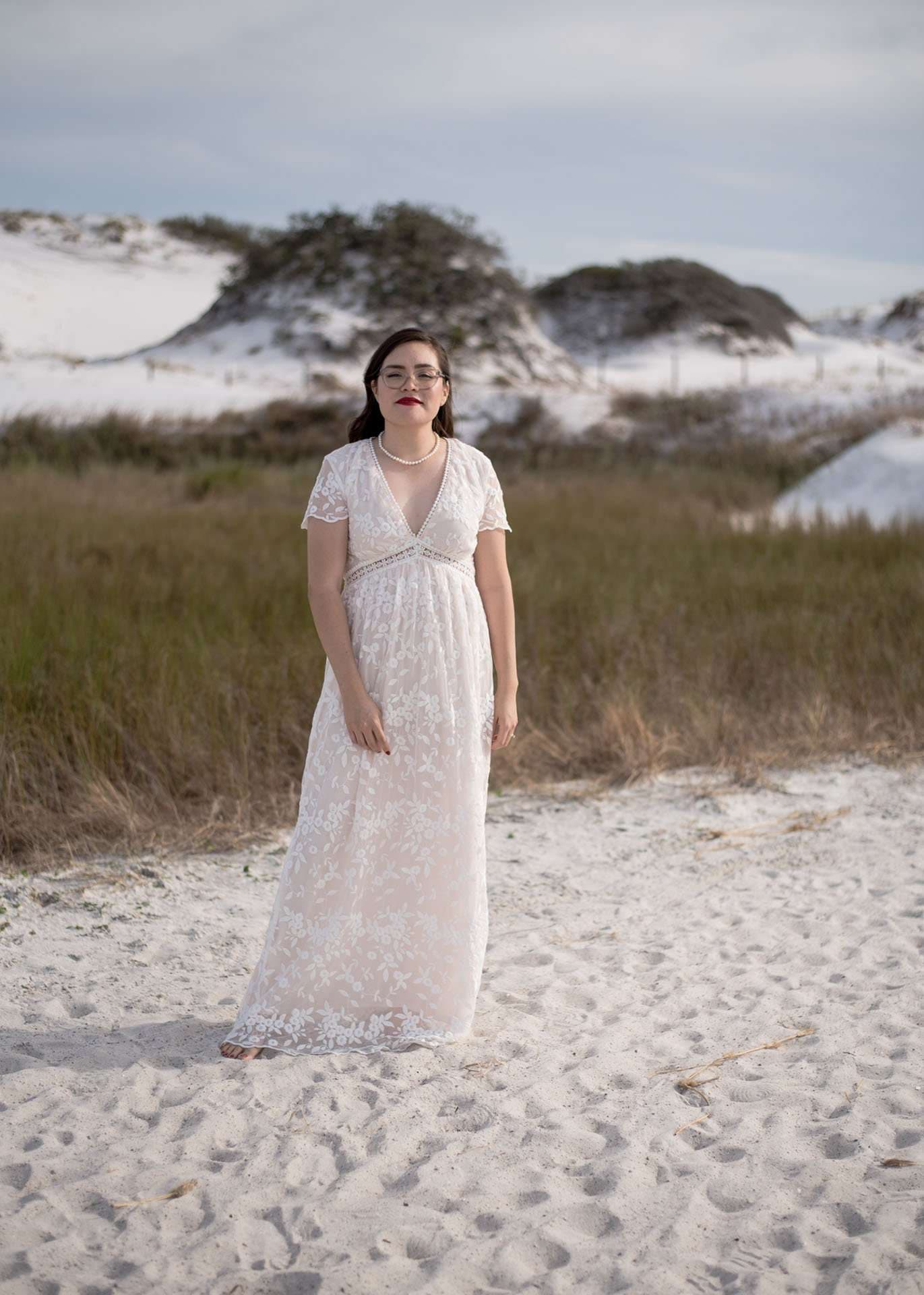 Woman in a white lace maxi gown and pearl necklace standing on white sand dunes with tall grass behind her