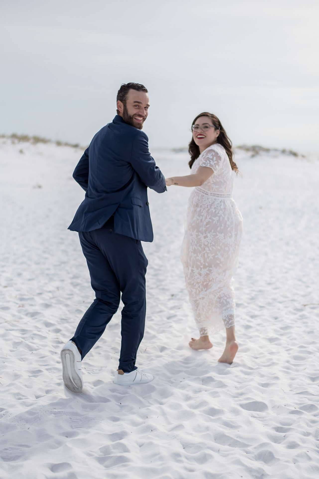 Groom in a navy suit leading his barefoot bride in a lace dress across white sand