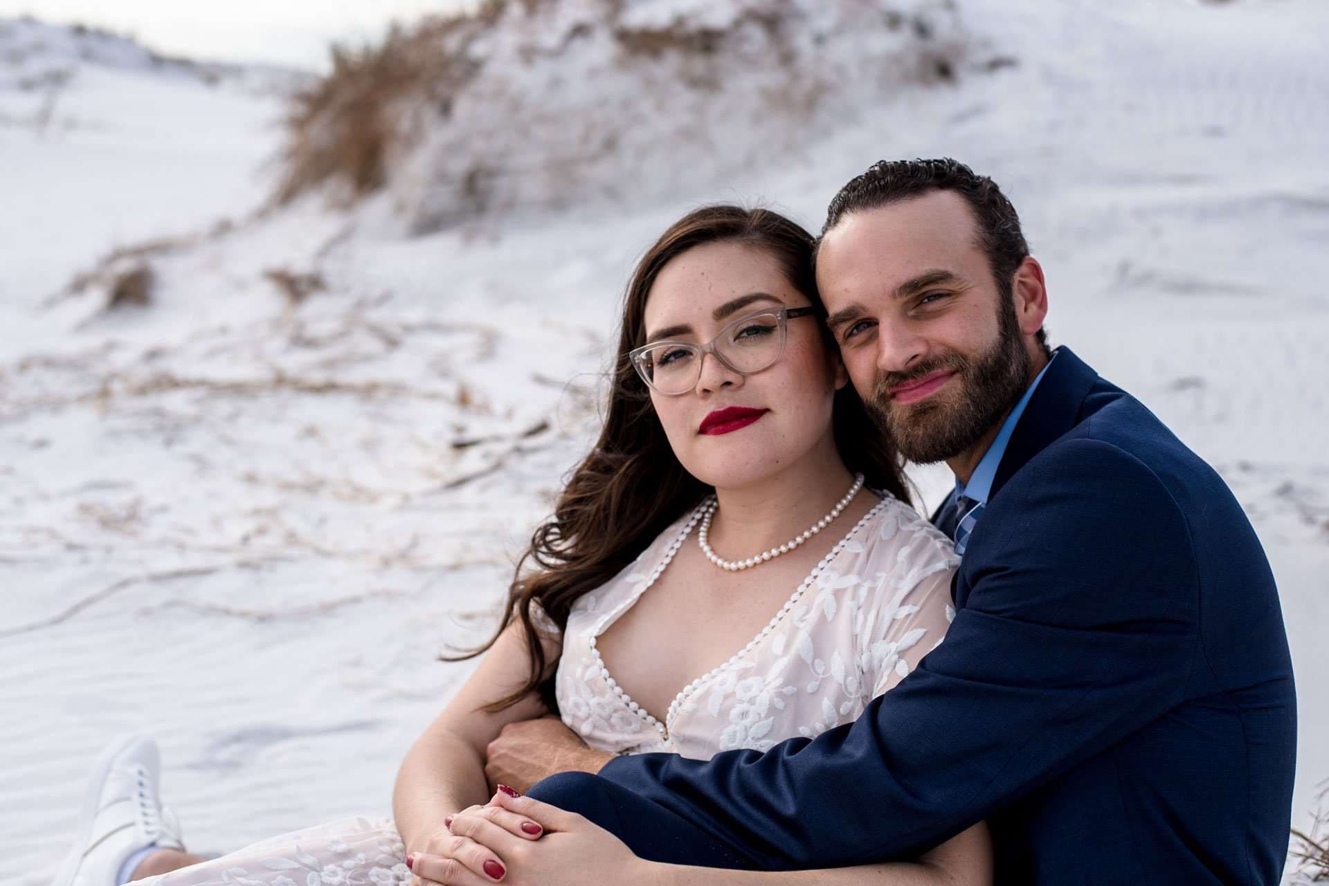 Smiling couple embracing on white sand dunes, woman in a lace dress and pearls, man in a navy suit