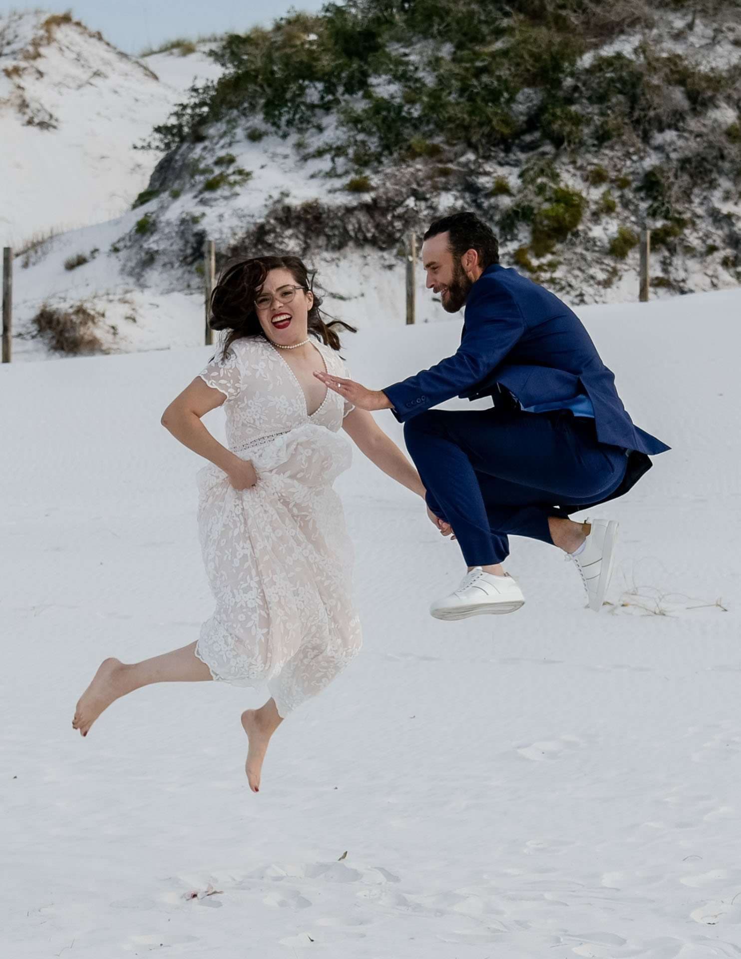 Couple in wedding attire leaping mid-air together on white sand dunes, she barefoot in a lace dress, he in a blue suit